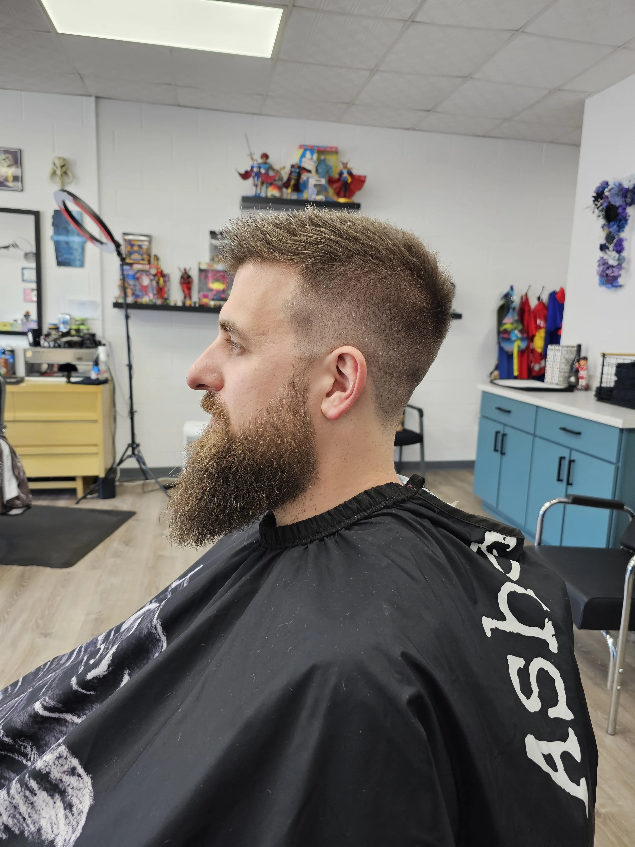 Side view of a young man with a fresh haircut, beard, and mustache sitting in a barber's chair in a barbershop. The background includes shelves with action figures and toys, a mirror, a yellow storage cabinet, and a blue cabinet.