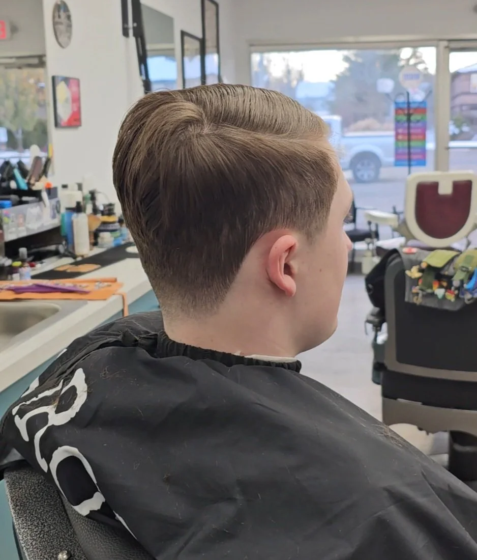 Side view of a young man with a freshly cut, styled haircut sitting in a barber shop, with hair clippings on his black barber cape, in a brightly lit salon with large windows showing an outside street scene.