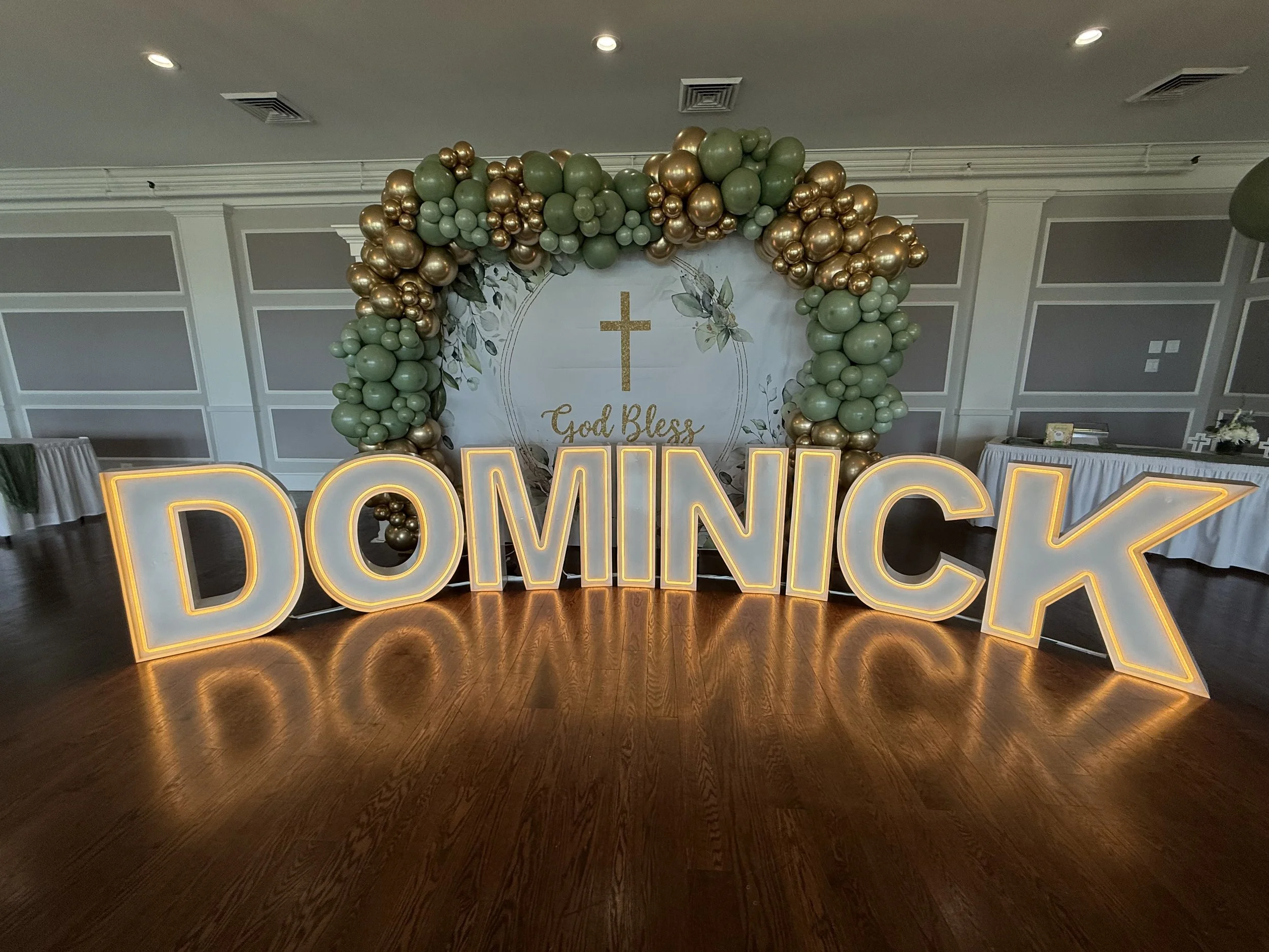 Large illuminated sign spelling 'DOMINICK' in front of decorative backdrop with balloons and a cross, featuring the words 'God Bless'.