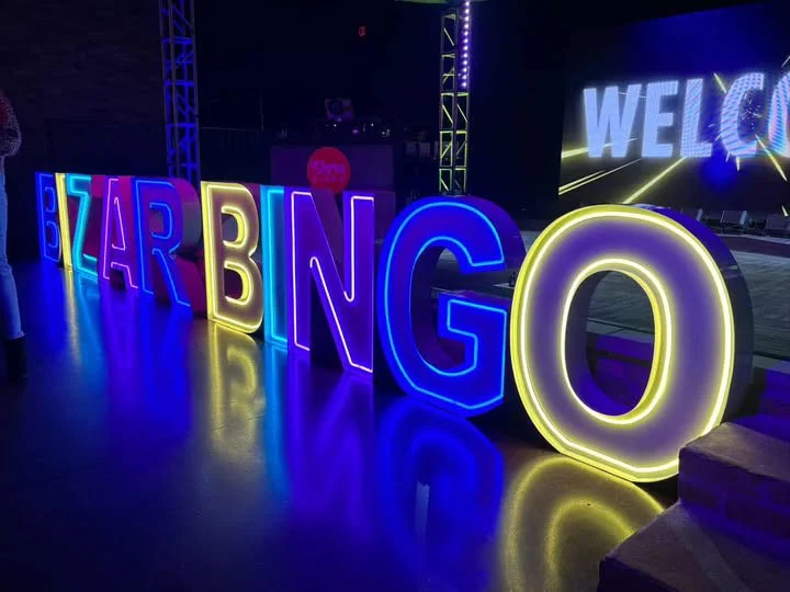 Colorful neon letters spell out "E-ZAR-BNGO," with a lit welcome screen in the background at an indoor event or party.