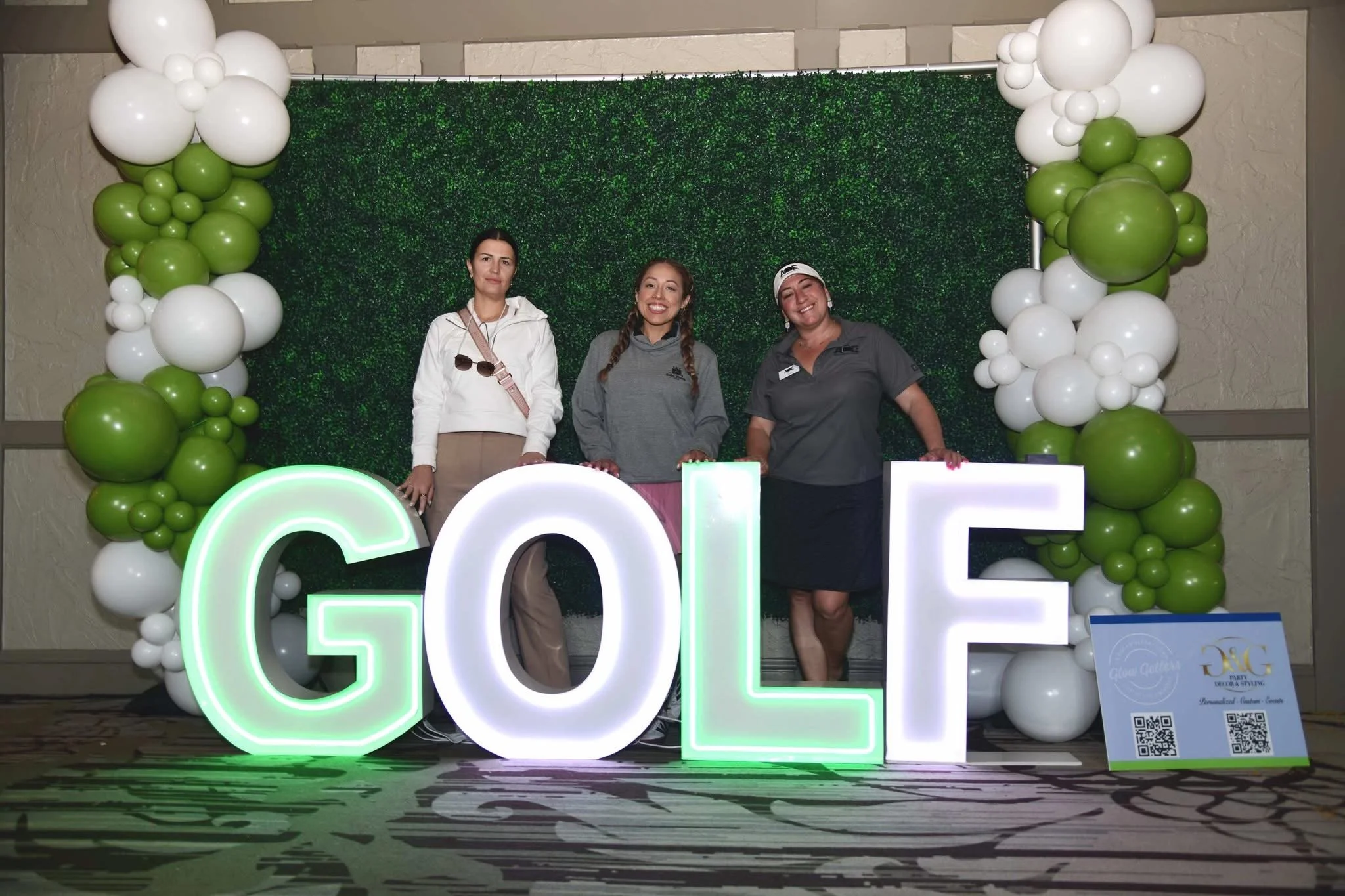 Three women standing behind large illuminated letters spelling 'GOLF' with balloon decorations in green and white and a green textured backdrop.