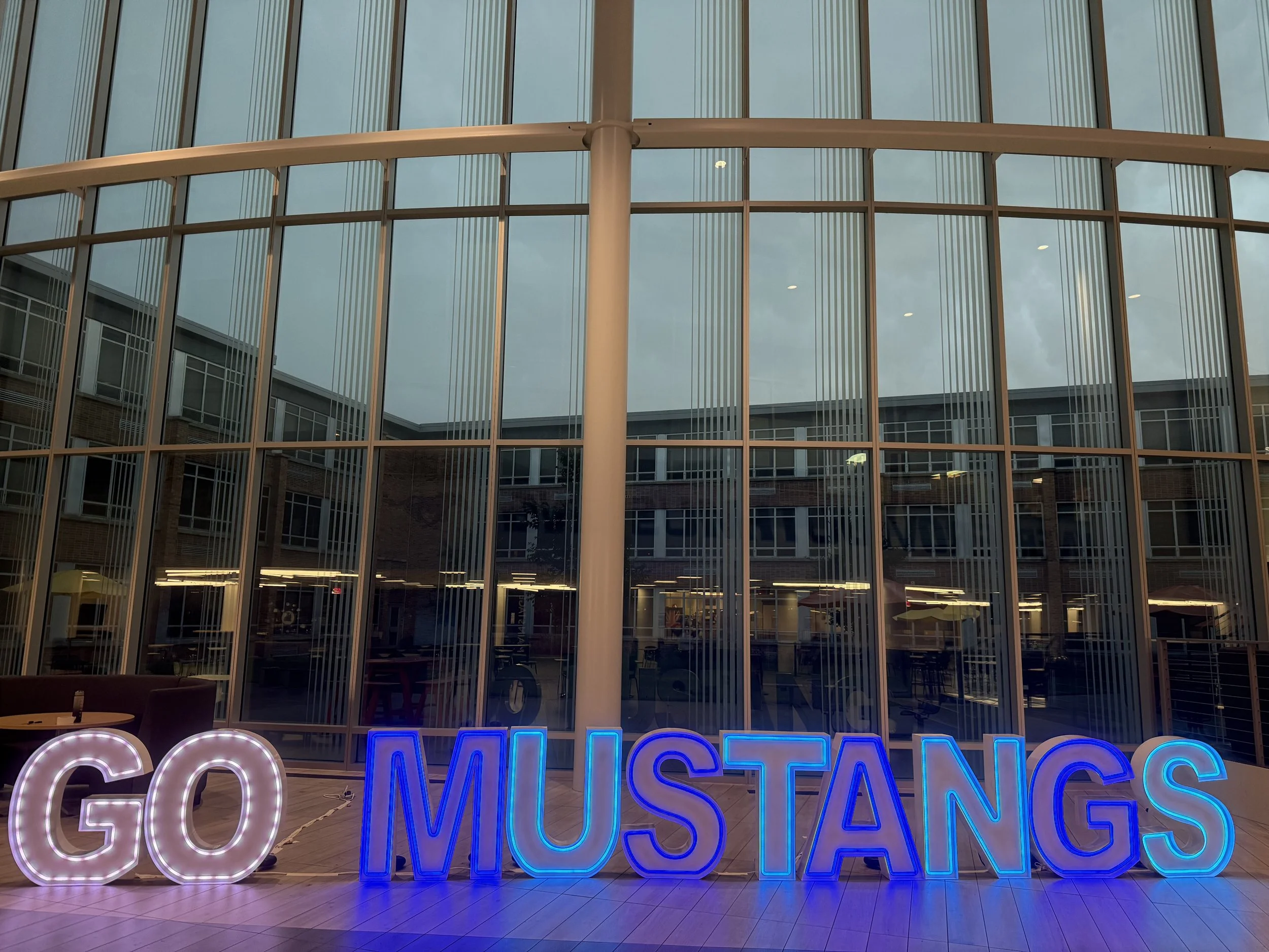 Neon sign spelling out 'GO MUSTANGS' in front of a glass windowed building.