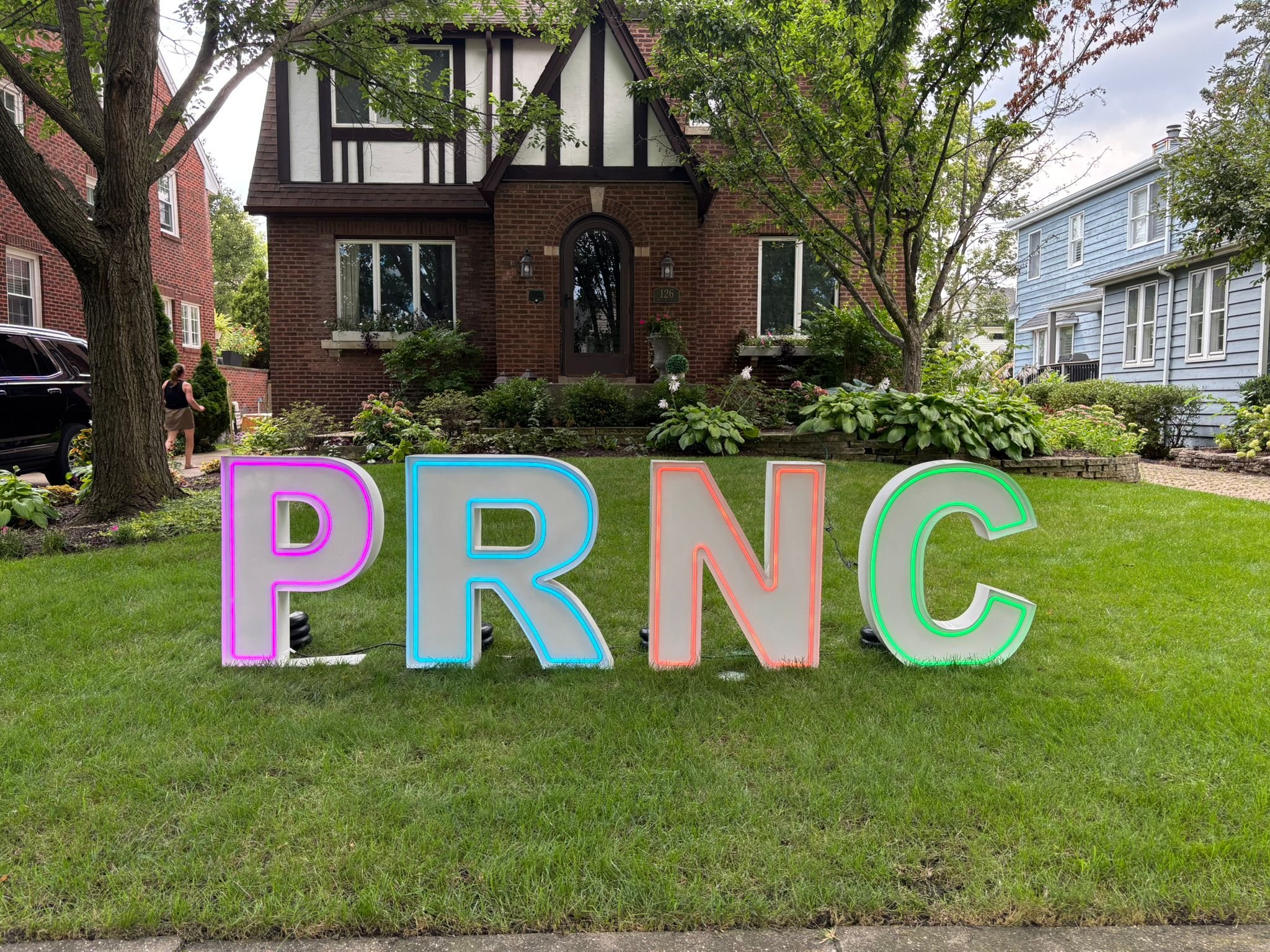 Colorful illuminated letters spelling 'PRNC' on a lawn in front of a brick house with trees and plants.