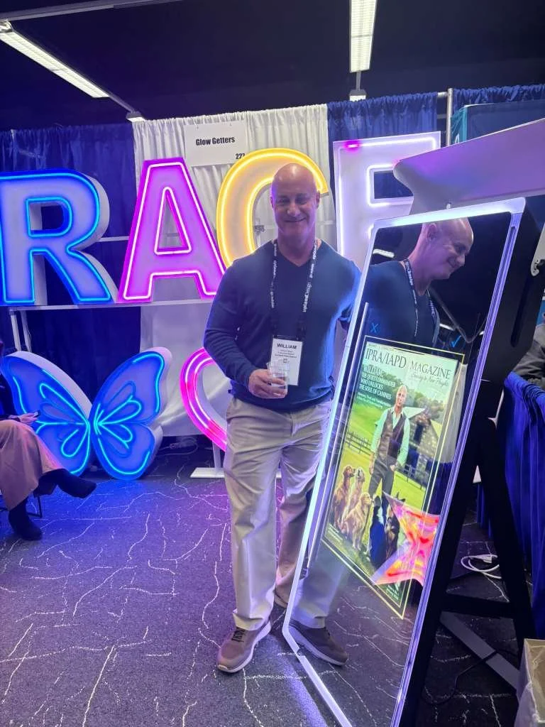 A man standing at a booth with neon lights spelling out 'RACE' and a butterfly-shaped neon sign. He's holding a glass and smiling. There is a magazine display in front of him showing a cover with a person and multiple dogs.