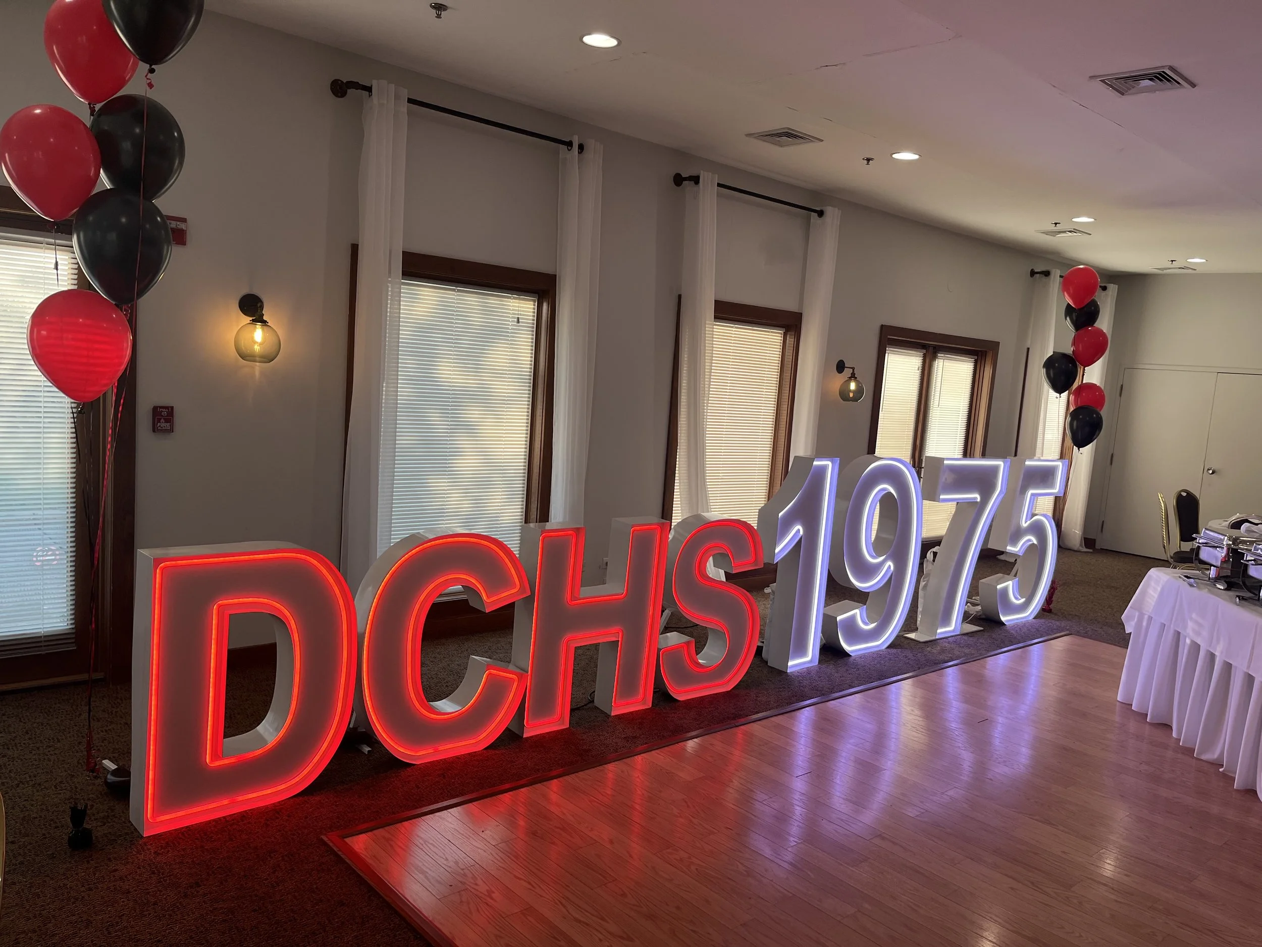 Decorative display with large illuminated letters reading 'DCHS 1975', flanked by black and red balloons, in a decorated event room with windows, white curtains, and a wooden floor.