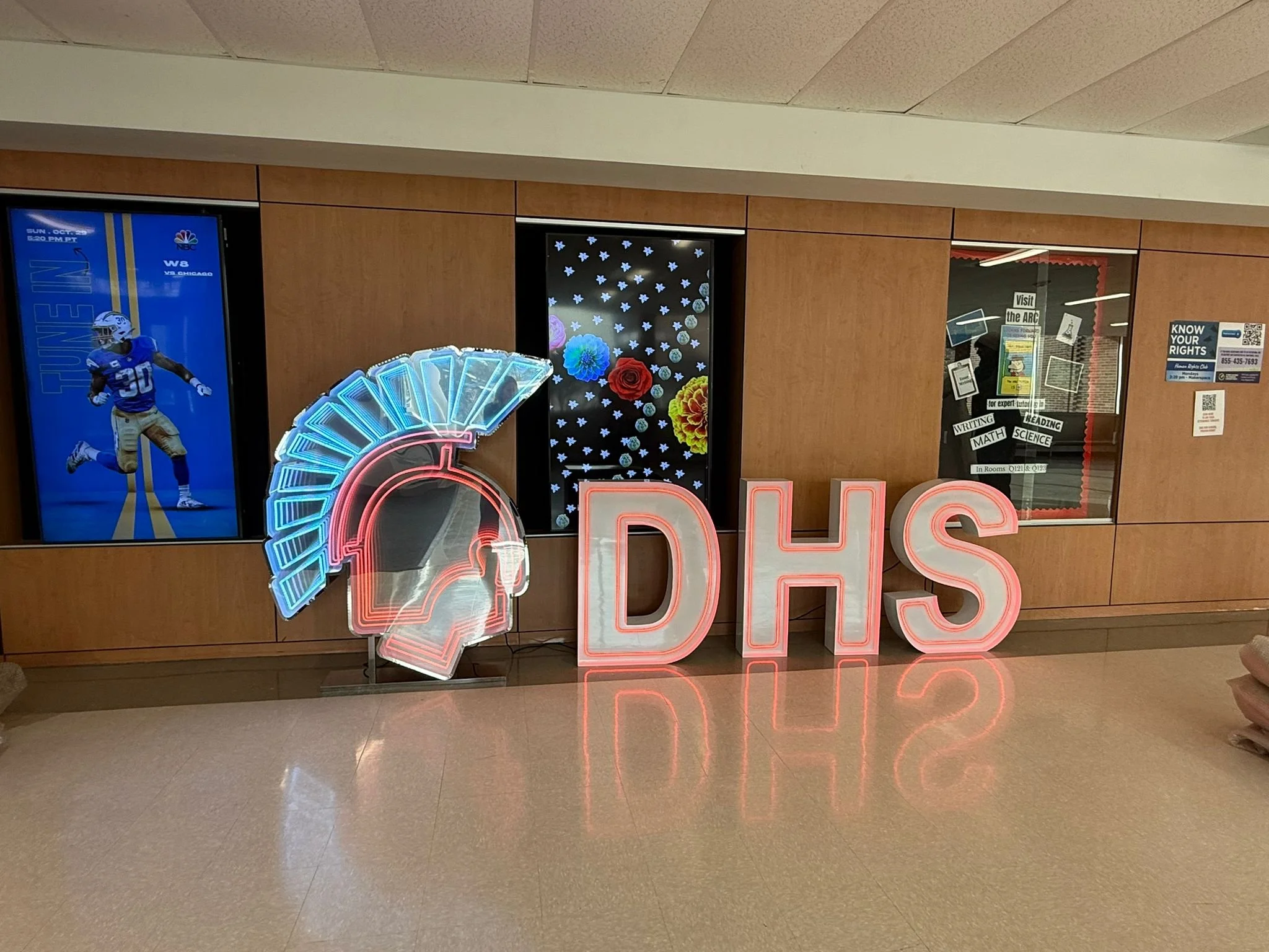 Neon sign of a football helmet and the letters 'DHS' in a school hallway. Behind the sign are wood-paneled walls with posters and windows, including one with a football player and sports information.