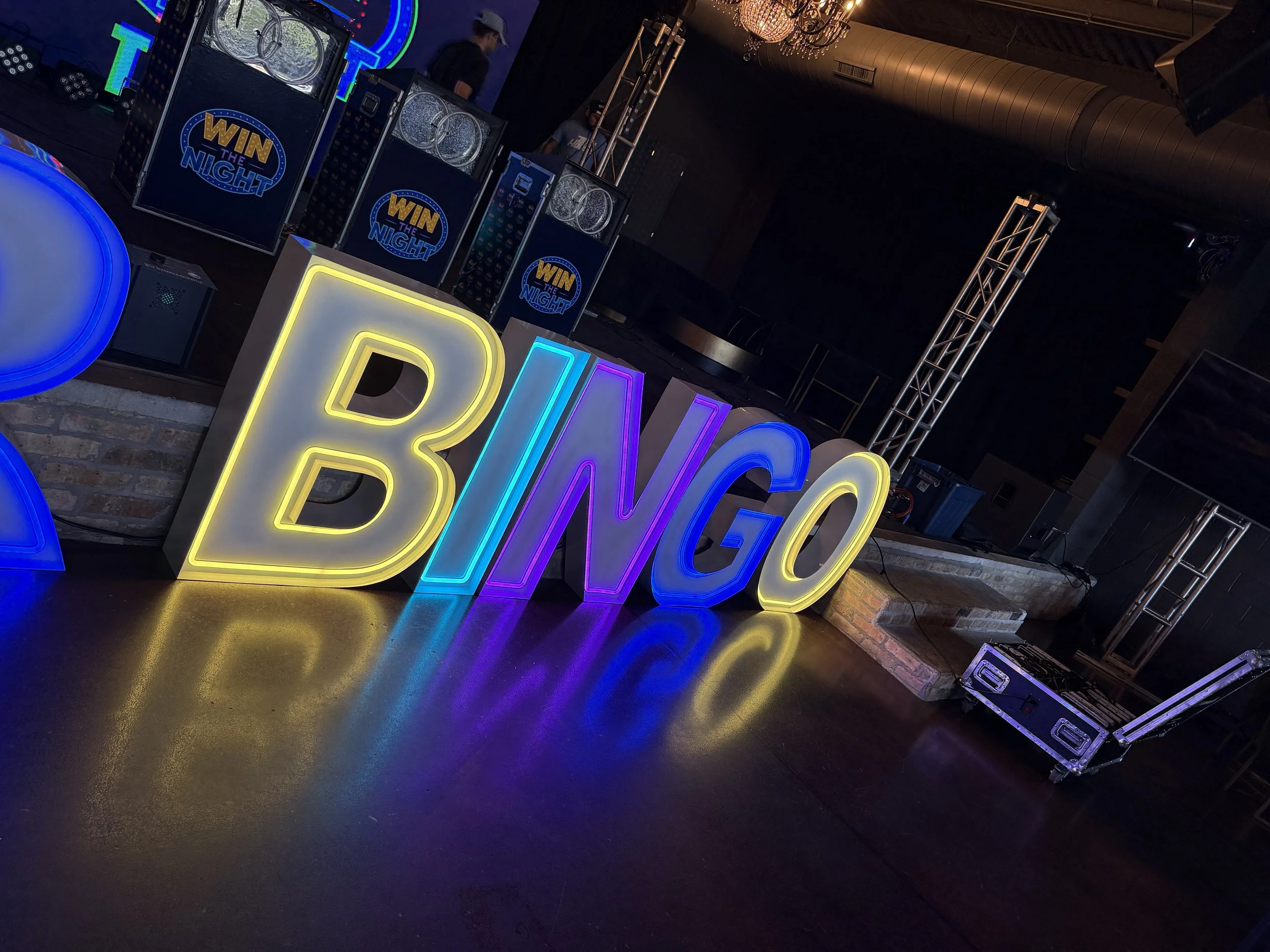 Colorful neon sign spelling BINGO on a dark party or event stage with DJ equipment, lighting, and people in the background.