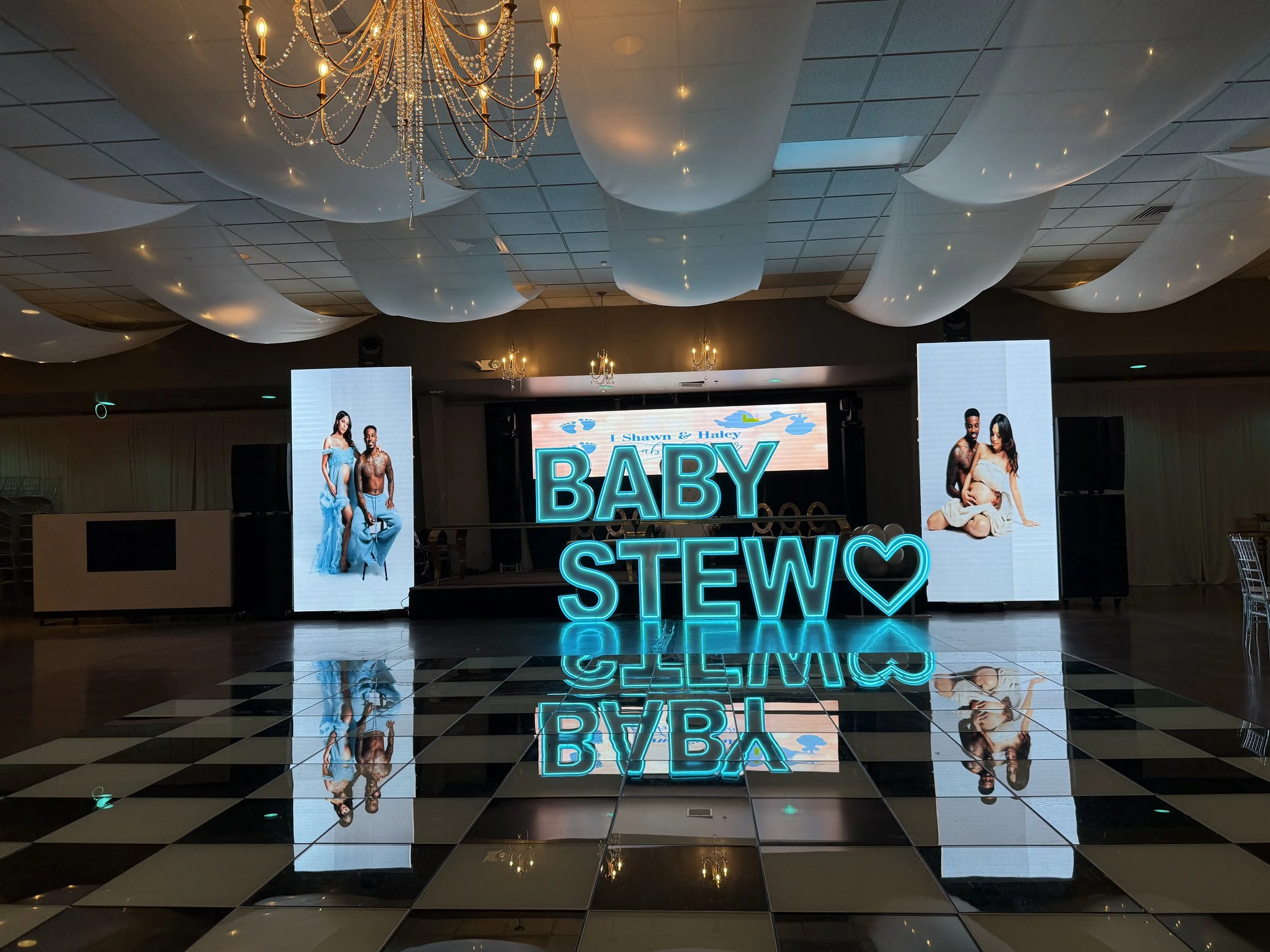 Decorated event hall with a black and white checkered floor, large illuminated sign that reads 'BABY STEW' with a heart symbol, two digital screens displaying a couple, and chandeliers hanging from the ceiling.