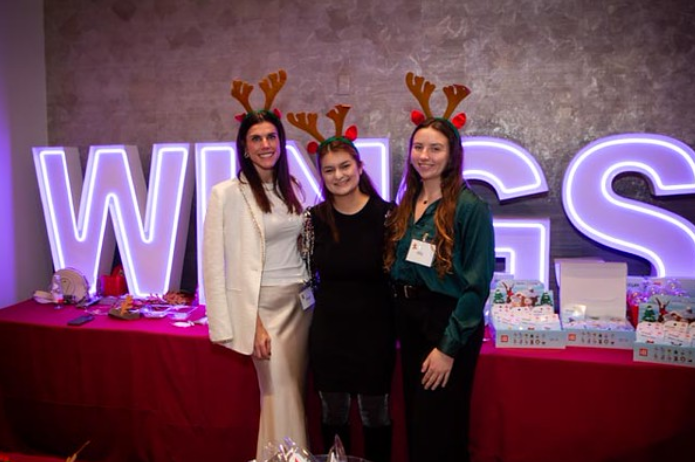 Three women wearing reindeer antler headbands posing in front of a large illuminated sign that spells WINGS with a table of gifts and treats behind them.