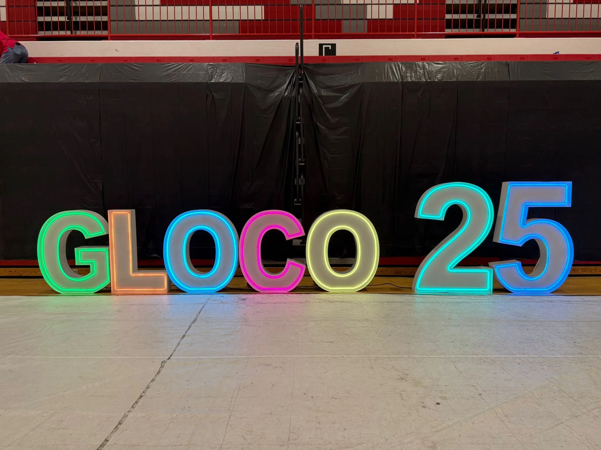 Colorful neon sign letters spelling 'GLOCO 25' on a gymnasium floor.