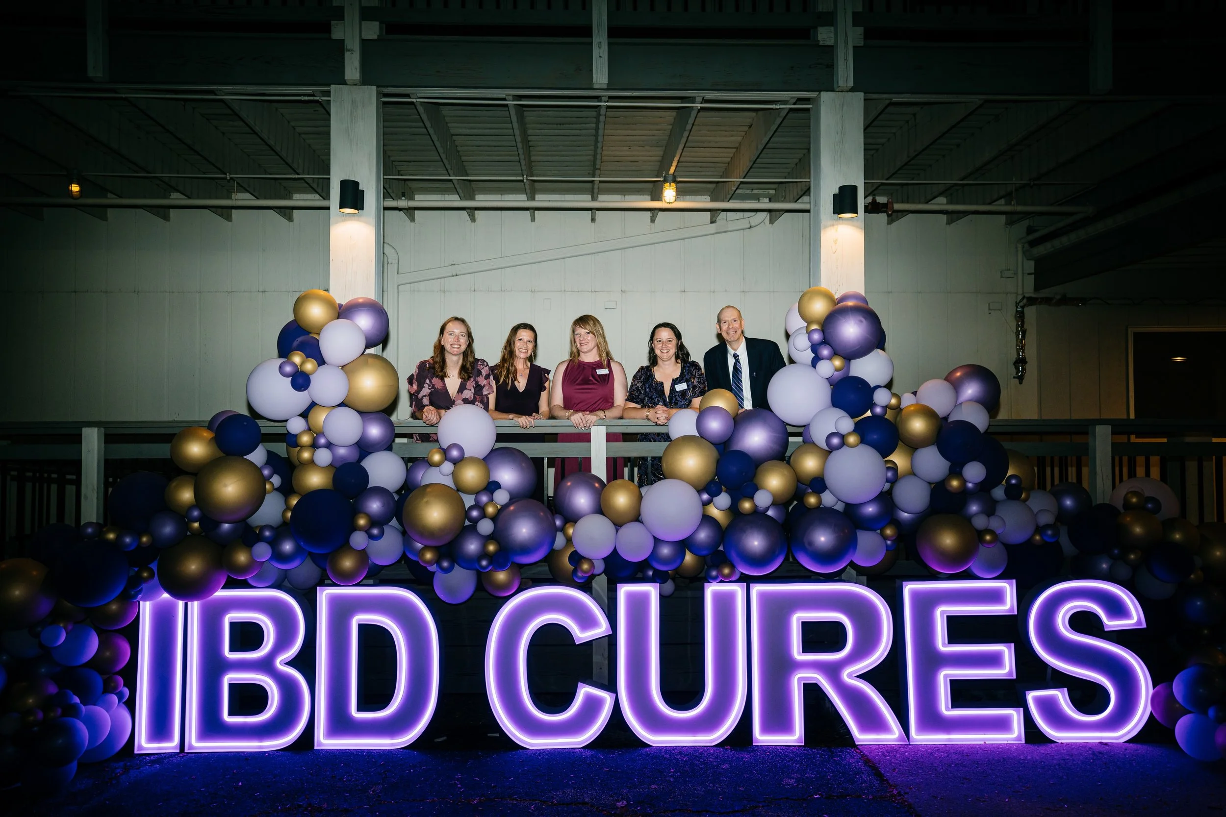 Group of five people standing behind a railing decorated with purple, white, gold, and navy balloons, with a neon sign that reads 'IBD CURES' in purple lights in front of them.
