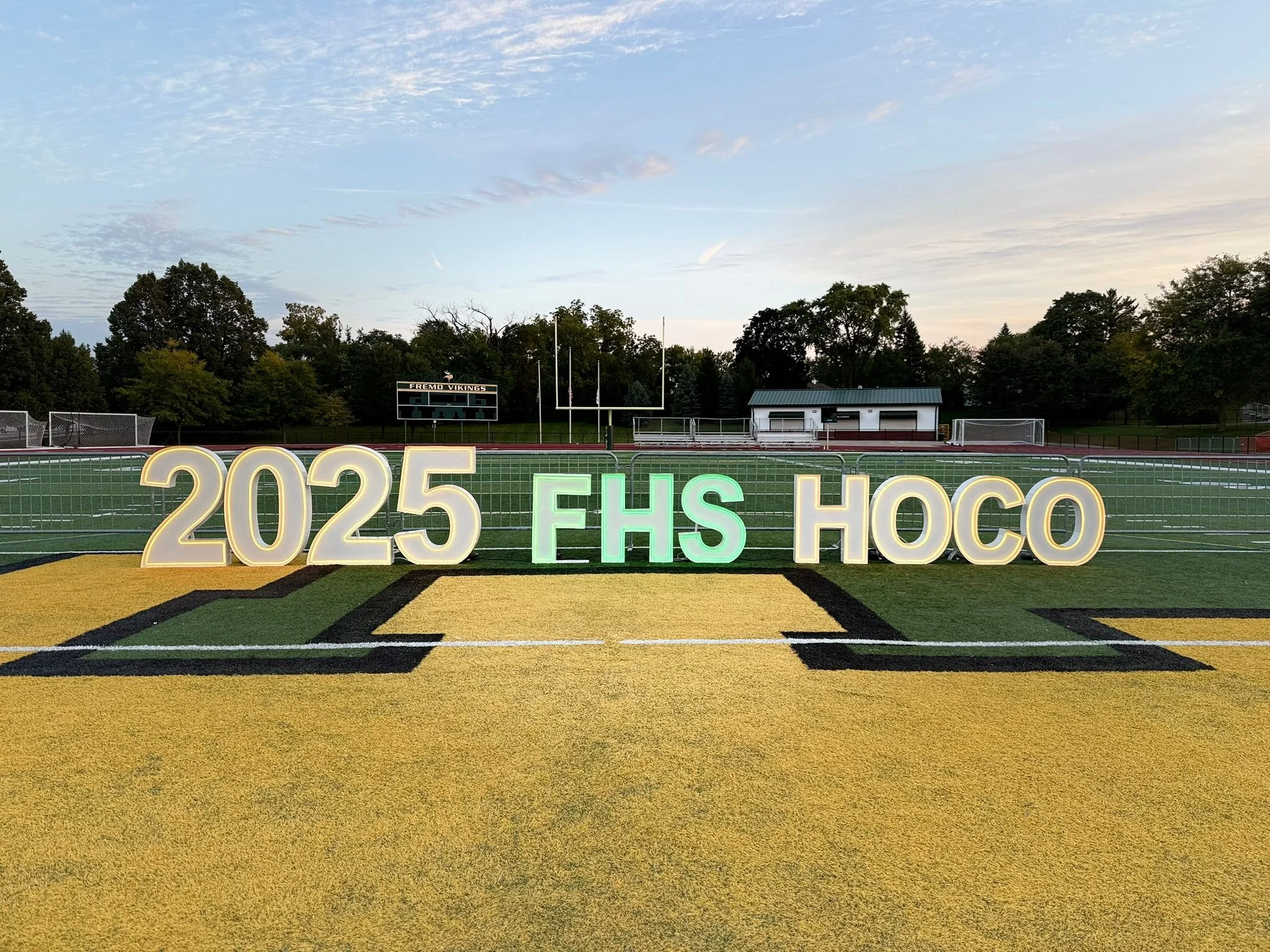 Football field with illuminated signs displaying "2025 EHS HOCO" at sunset, with trees, a scoreboard, and goalposts in the background.