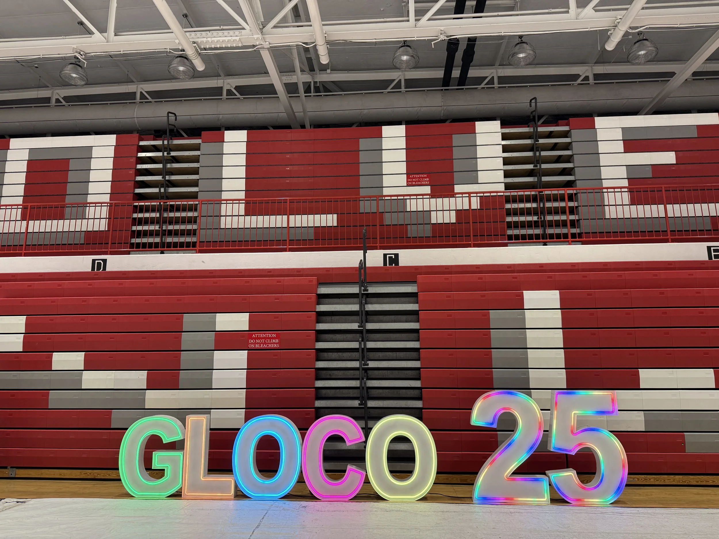Colorful illuminated letters spelling 'GLOCO 25' placed on a gymnasium floor in front of red, white, and gray stadium seating and bleachers.