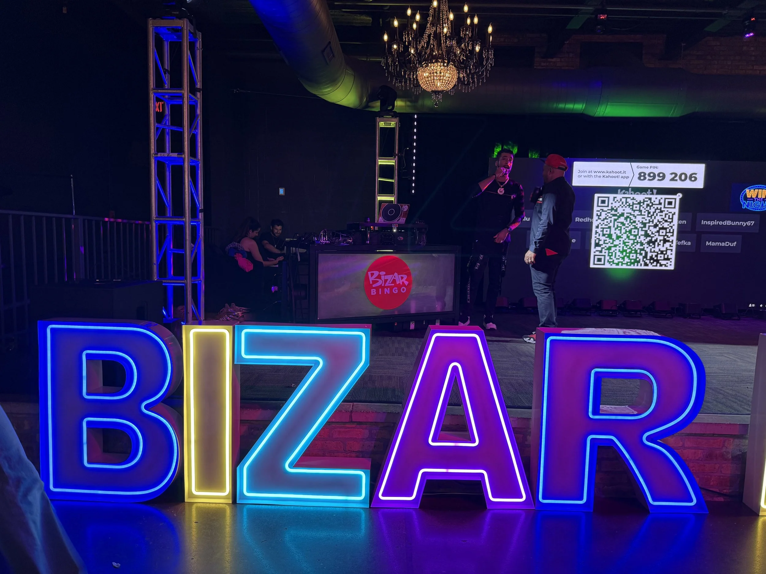 Colorful neon sign spelling 'BIZAR' in front of a stage with people and a DJ booth, event space with chandeliers and a large screen displaying a QR code and game PIN.
