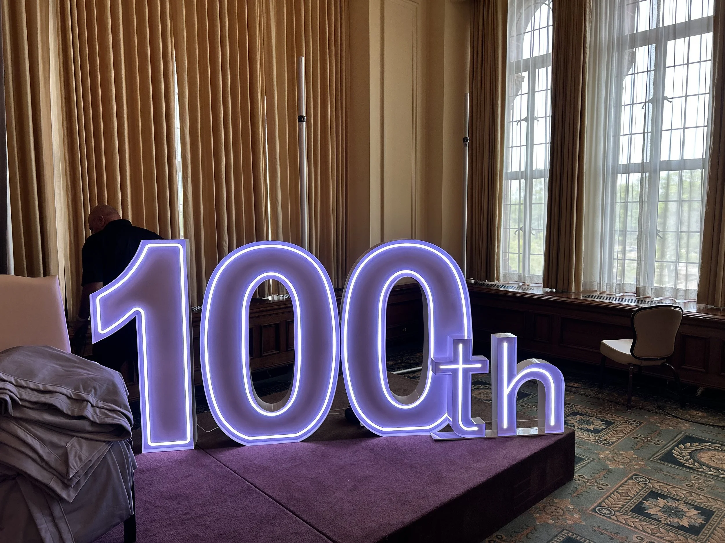 Large illuminated sign displaying '100th' in a room with wooden paneling, tall windows with curtains, and a patterned rug.