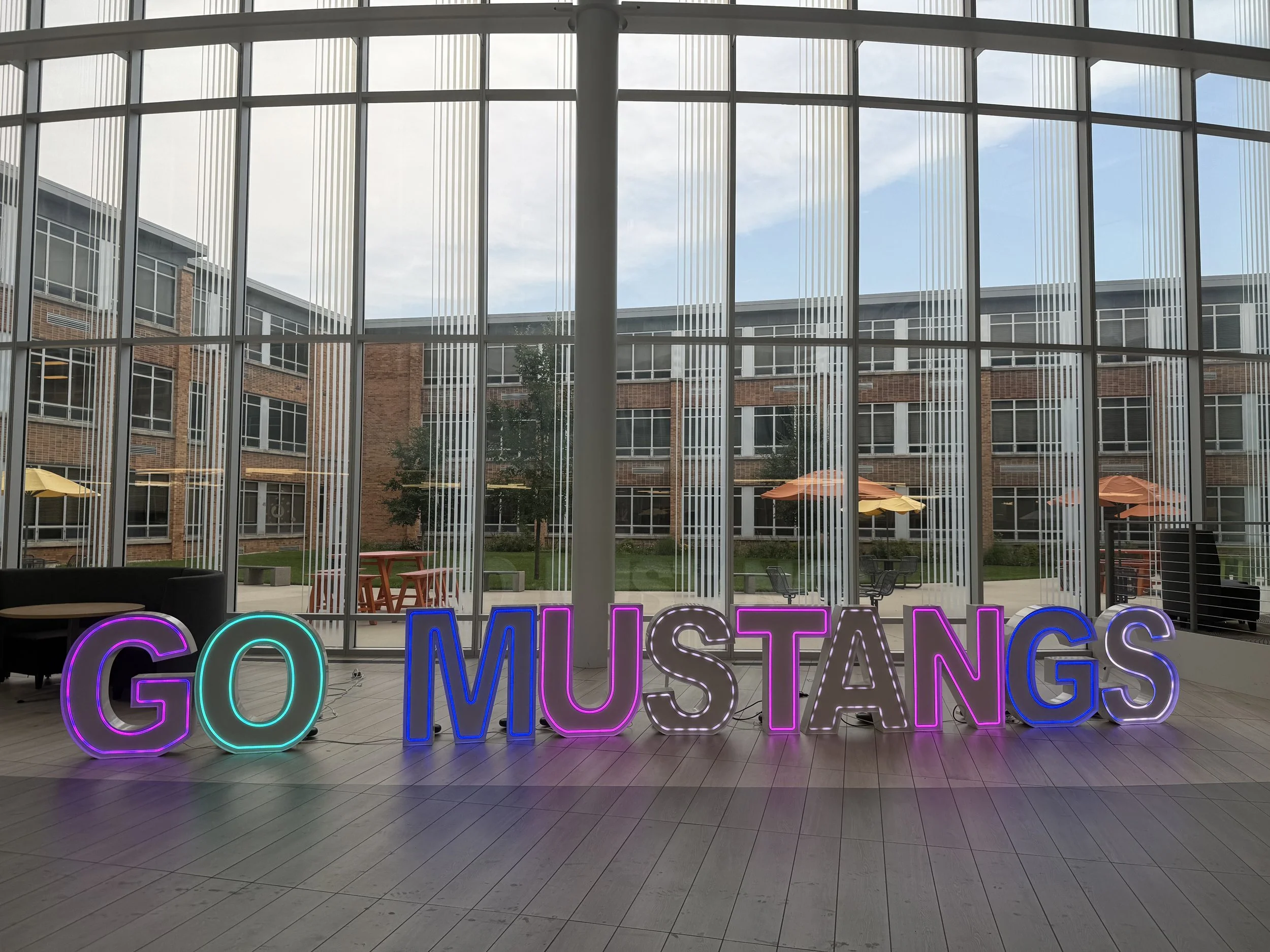 Neon sign reading 'GO MUSTANGS' with a large 'G' in purple and green, 'O' in green, 'MUSTANGS' in pink and blue, inside a modern glass building with windows showing an outdoor courtyard with tables, umbrellas, trees, and benches.