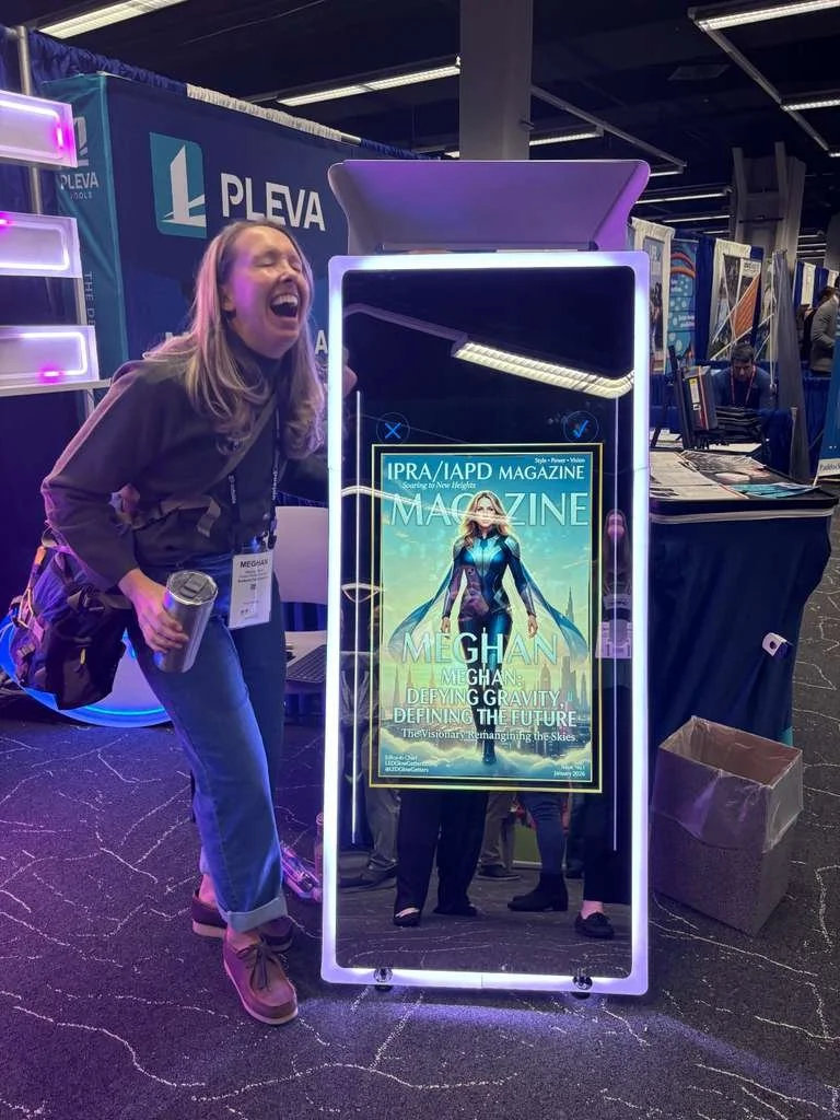 A woman standing next to a large mirror display at a convention, with a magazine cover featuring a superhero costume and the name Meghan on the mirror, as she laughs and holds a water bottle.