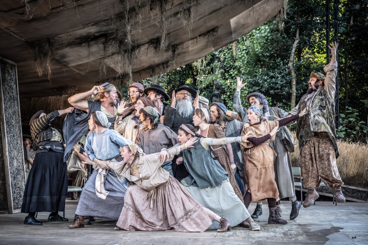 Group of actors performing on stage with dramatic expressions, wearing rustic, folk-inspired costumes, surrounded by a natural outdoor setting.