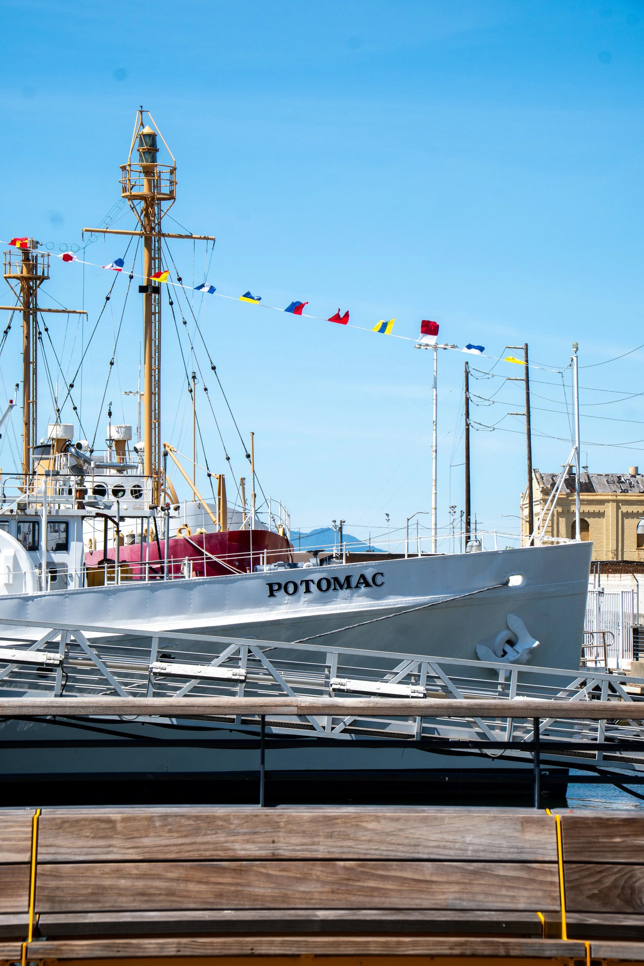 A docked ship named Potomac with a white hull, yellow masts, and colorful flags against a bright blue sky, with some buildings and utility poles in the background.