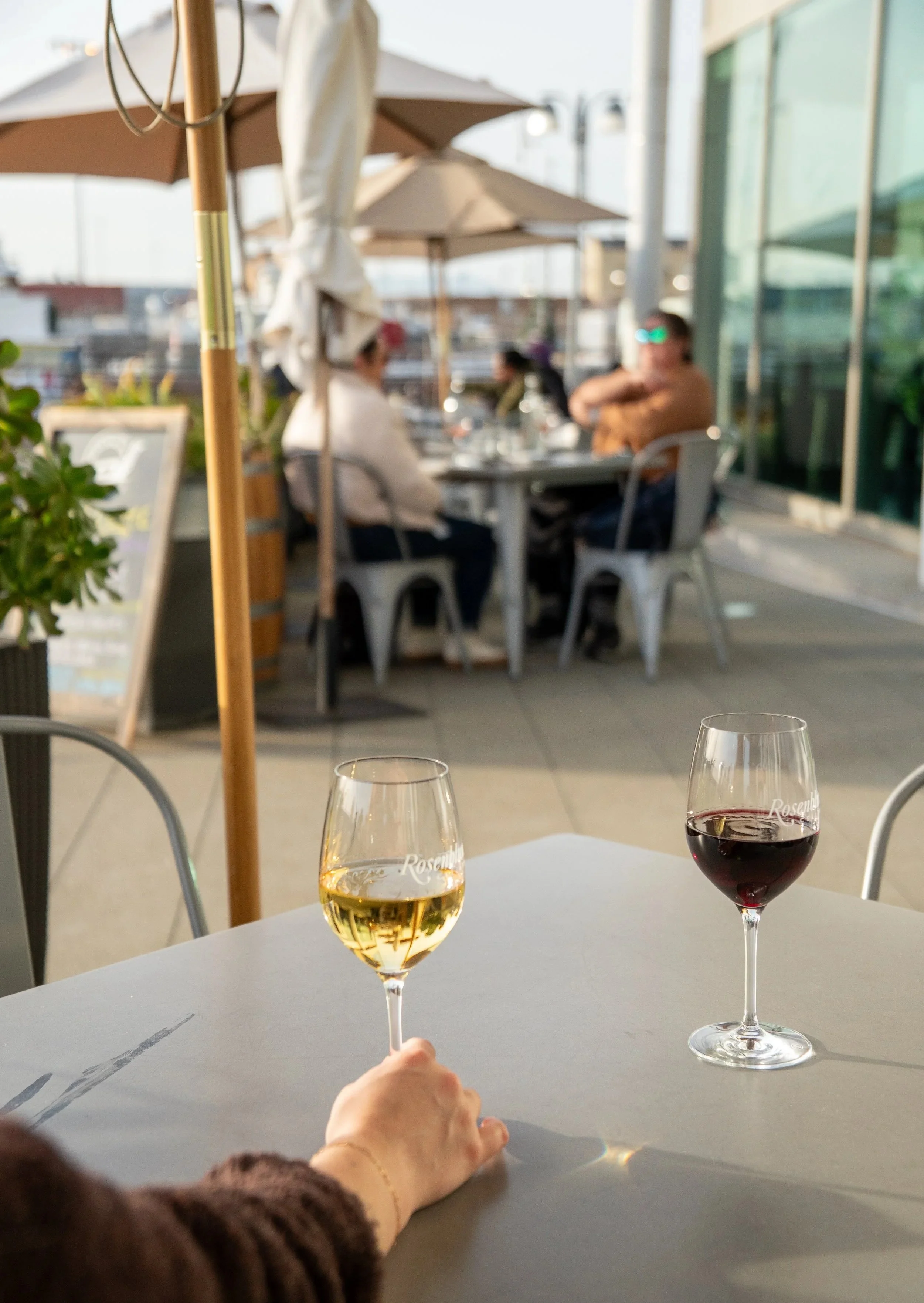 A table with two glasses of wine, one white and one red. In the background, there are people sitting under umbrellas at an outdoor dining area, with blurred buildings and glass windows.