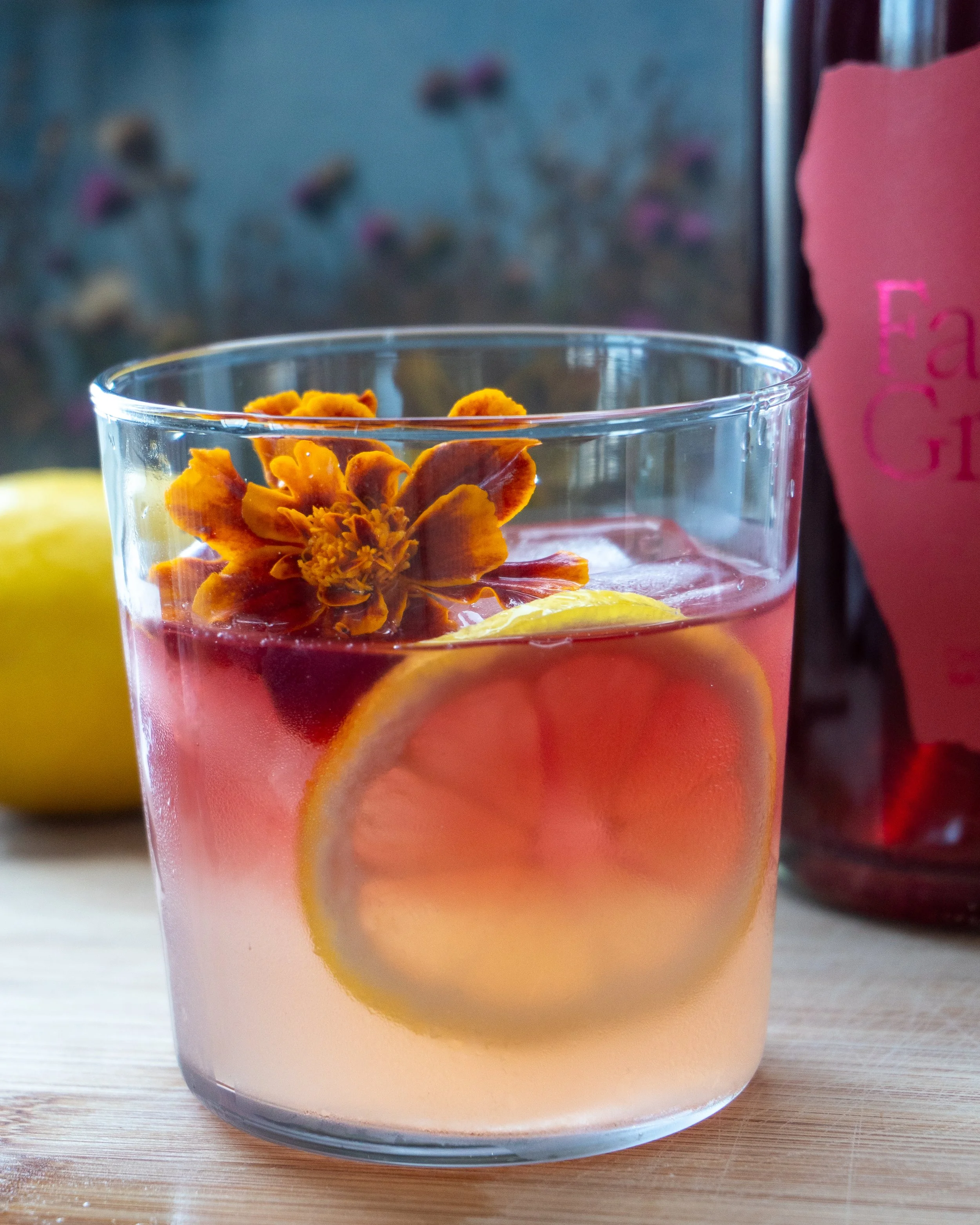 Close-up of a glass of pink drink with slices of lemon and an orange flower floating on top, placed on a light wood surface, with blurred background including a pink bottle of wine and a lemon