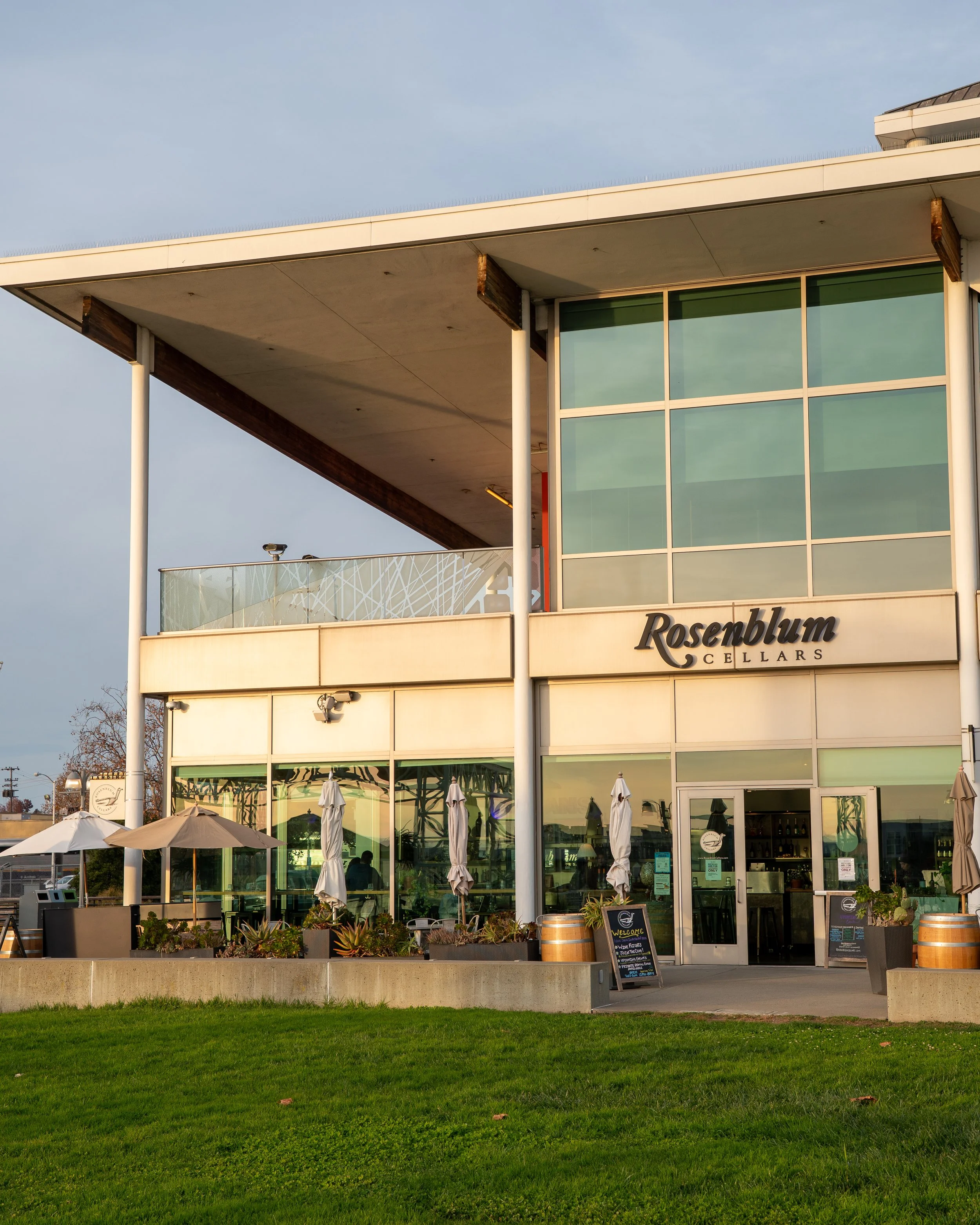 Exterior view of Rosenblum Cellars building with large glass windows, outdoor patio with umbrellas, and a well-maintained lawn in front.