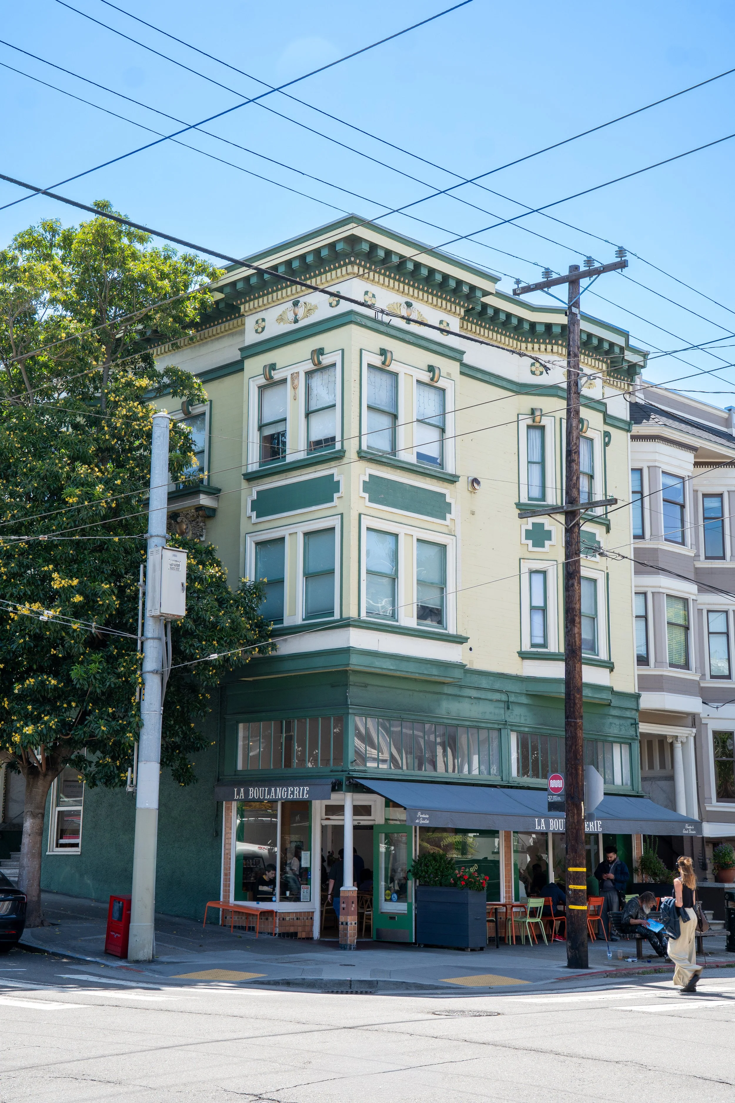 A corner building with a yellow and green facade, housing a bakery called La Boulangerie, with outdoor seating, trees, people walking and sitting, and power lines overhead on a sunny day.