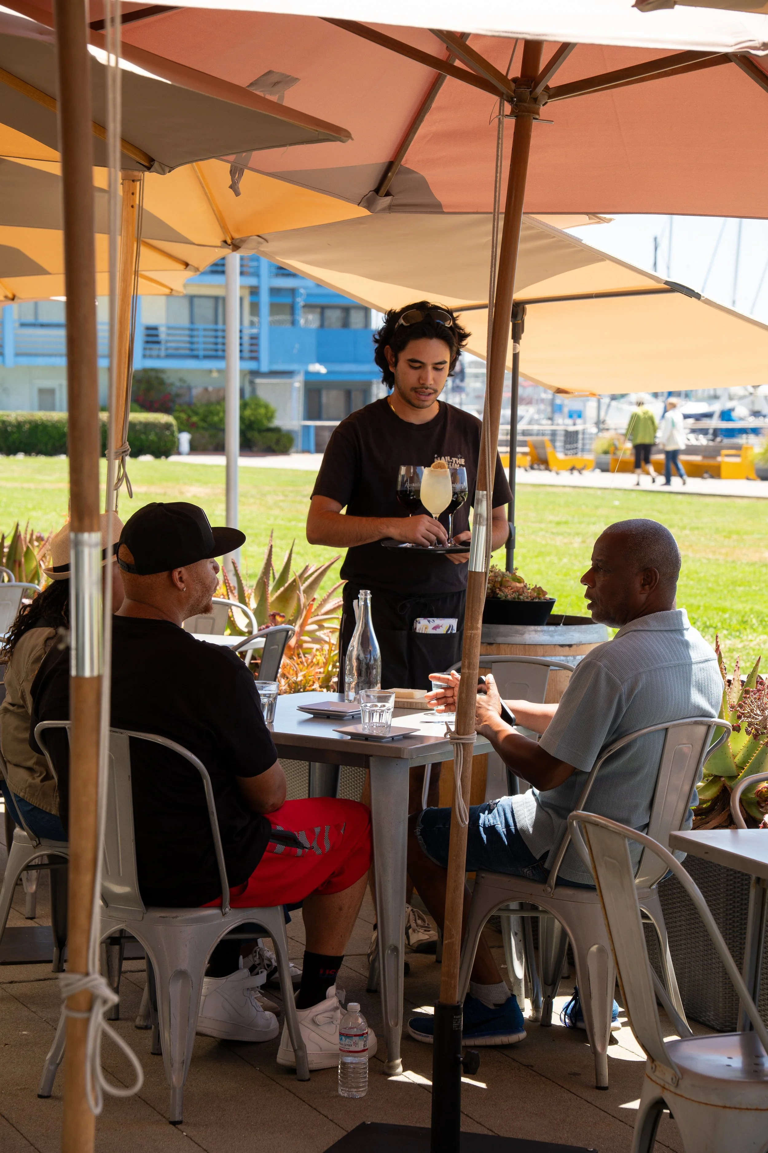 A group of people seated at an outdoor restaurant table under large umbrellas on a sunny day. A waiter is serving drinks to two people at the table, one in a white shirt and the other in a black shirt and red shorts.