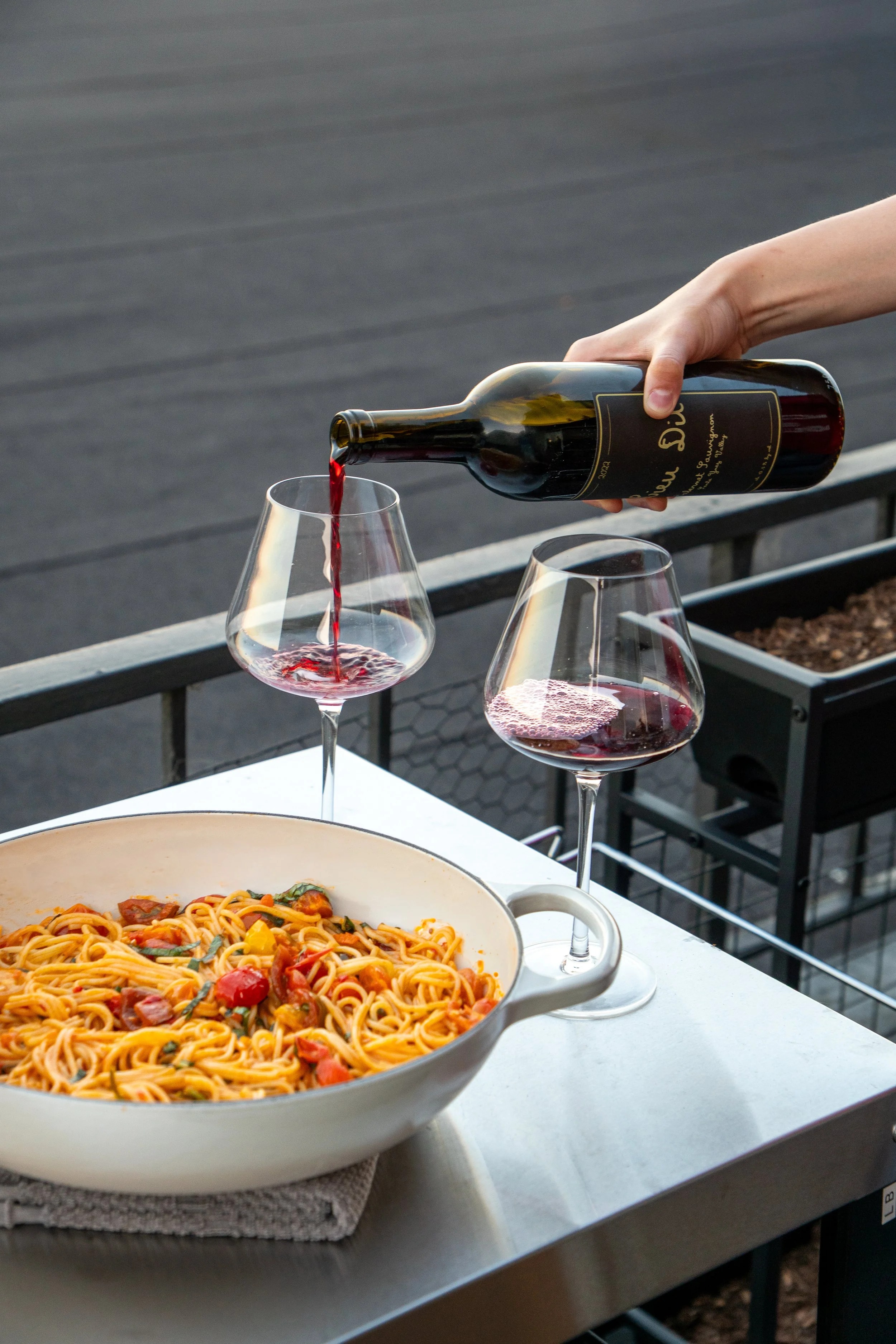 A person pouring red wine into two glasses on an outdoor table, with a bowl of spaghetti with tomatoes in the foreground and a rooftop in the background.