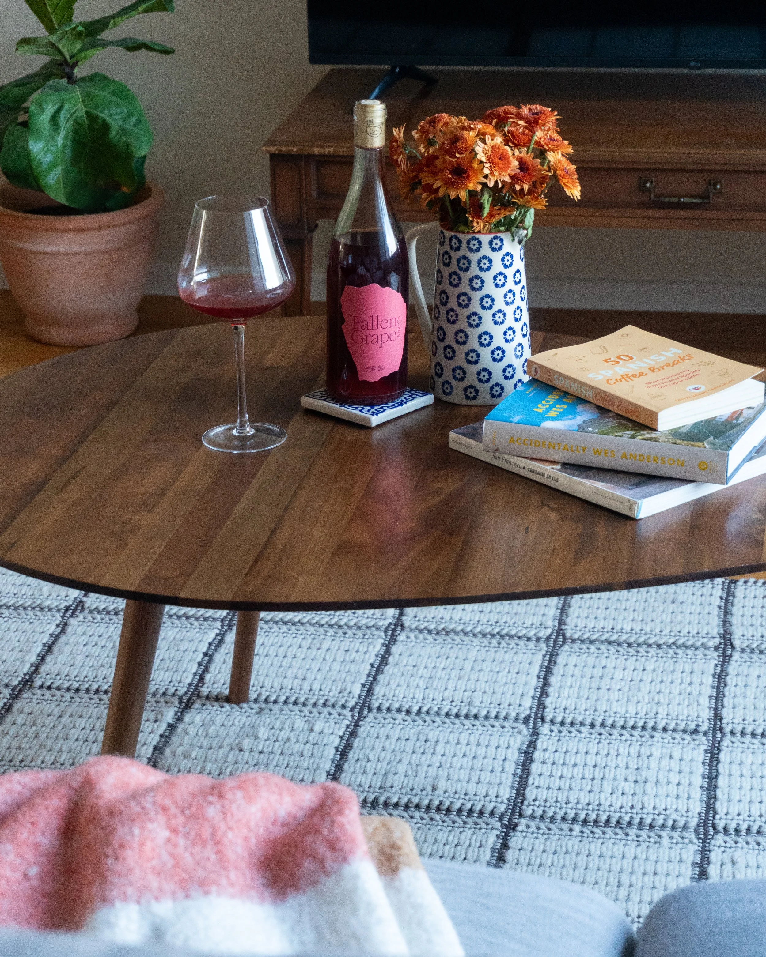 A wooden table with a glass of red wine, a bottle of red wine labeled 'Fallen Grape', a mug of orange and yellow flowers, and a stack of three books, with a large green potted plant and a wall-mounted TV in the background.