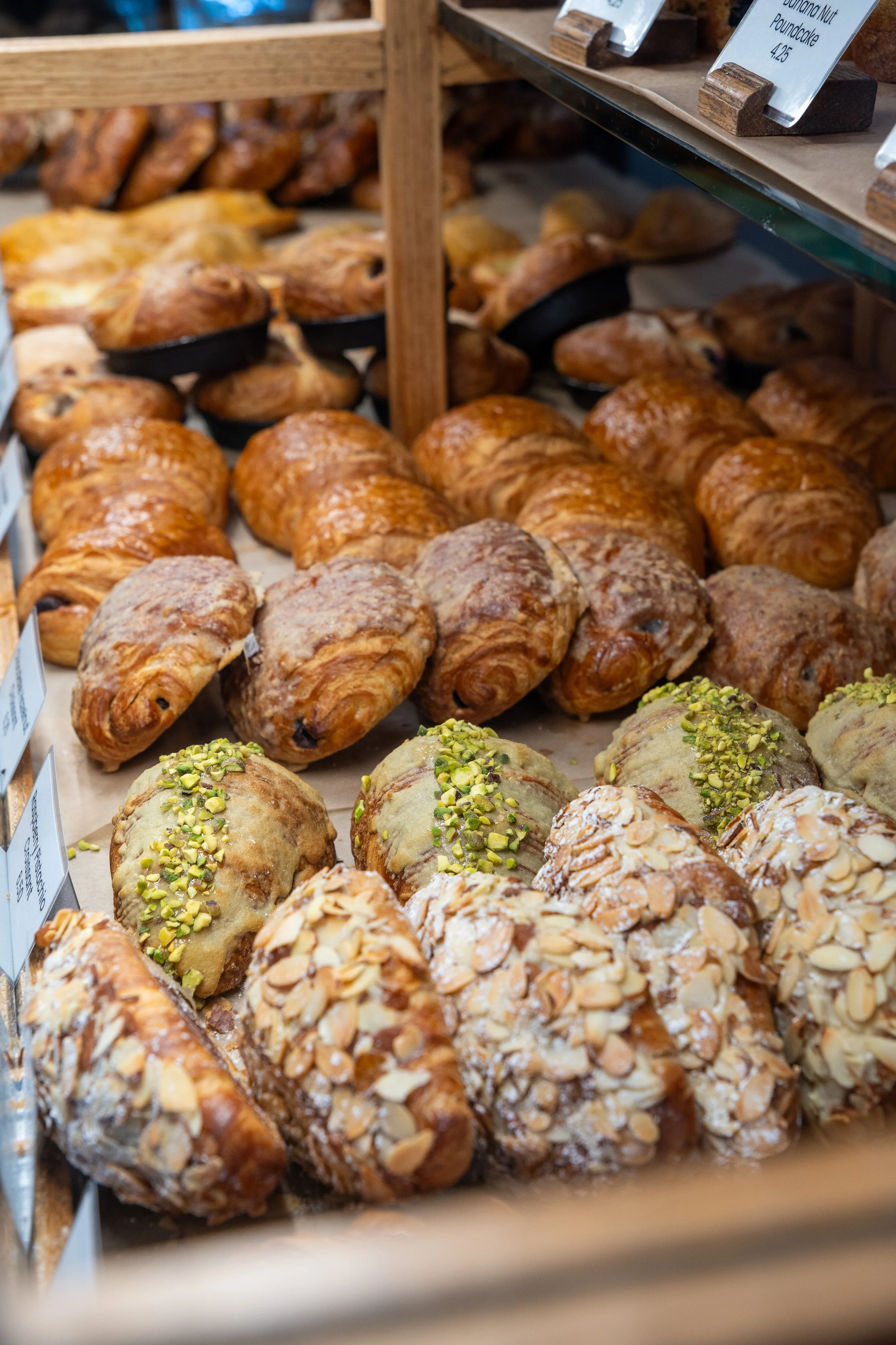 Display of assorted pastries, including croissants topped with chopped pistachios and other baked goods, on a wooden shelf in a bakery.