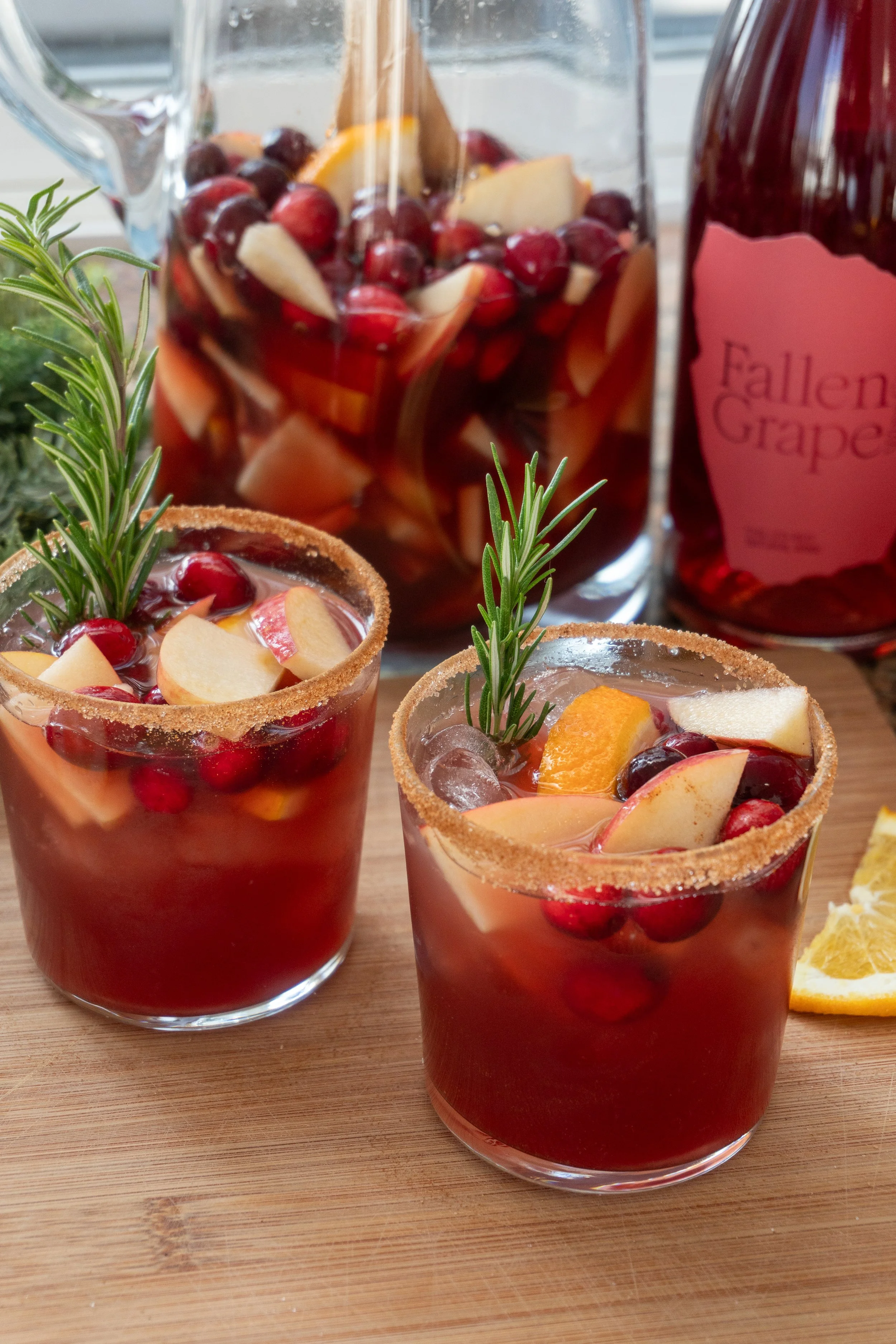 Three glasses of red sangria with assorted fruit pieces, garnished with sprigs of rosemary on a wooden surface, with a bottle labeled 'Fallen Grape' in the background.