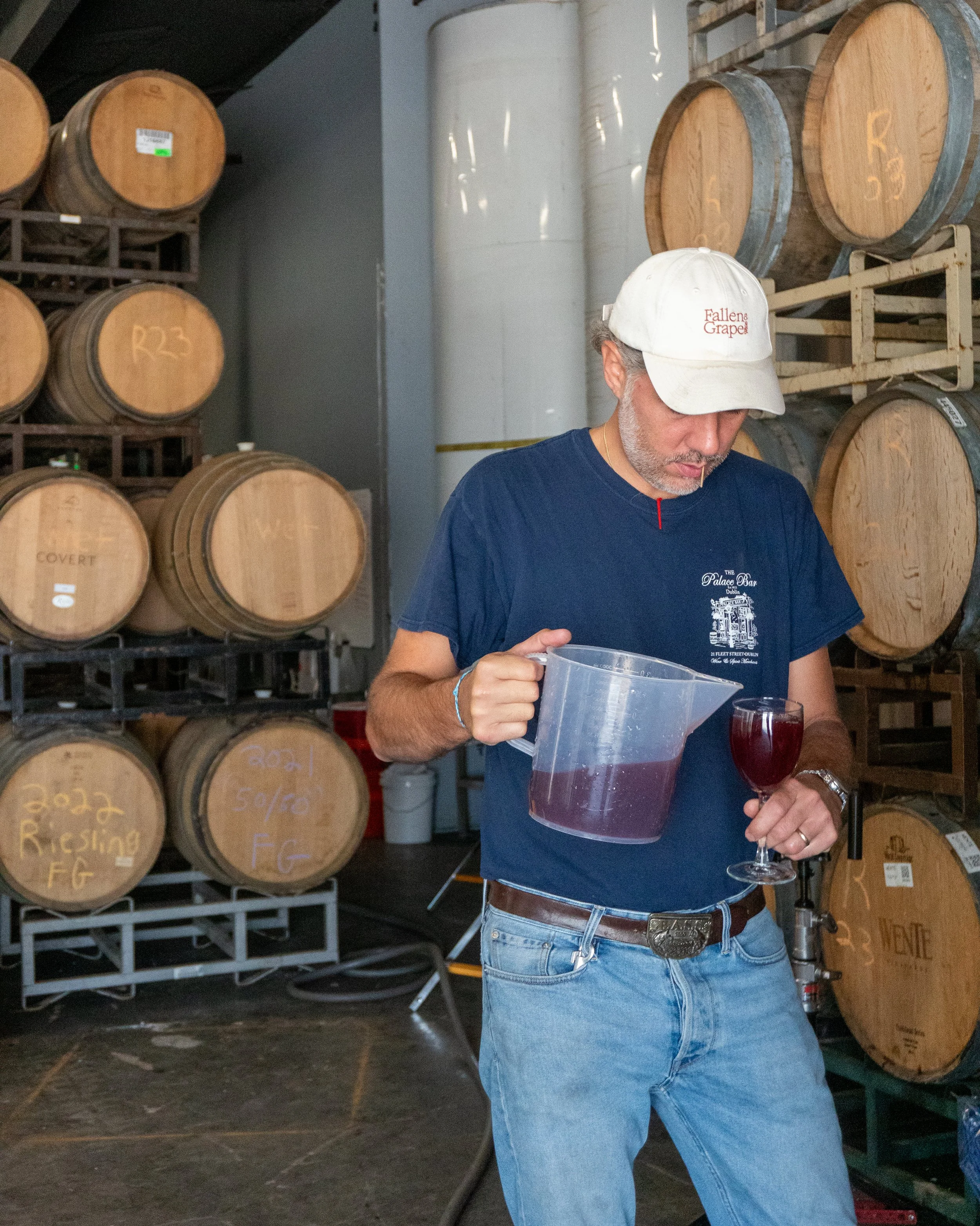 A man in a white cap and blue t-shirt standing near stacks of wooden wine barrels in a cellar, holding a glass of red wine.