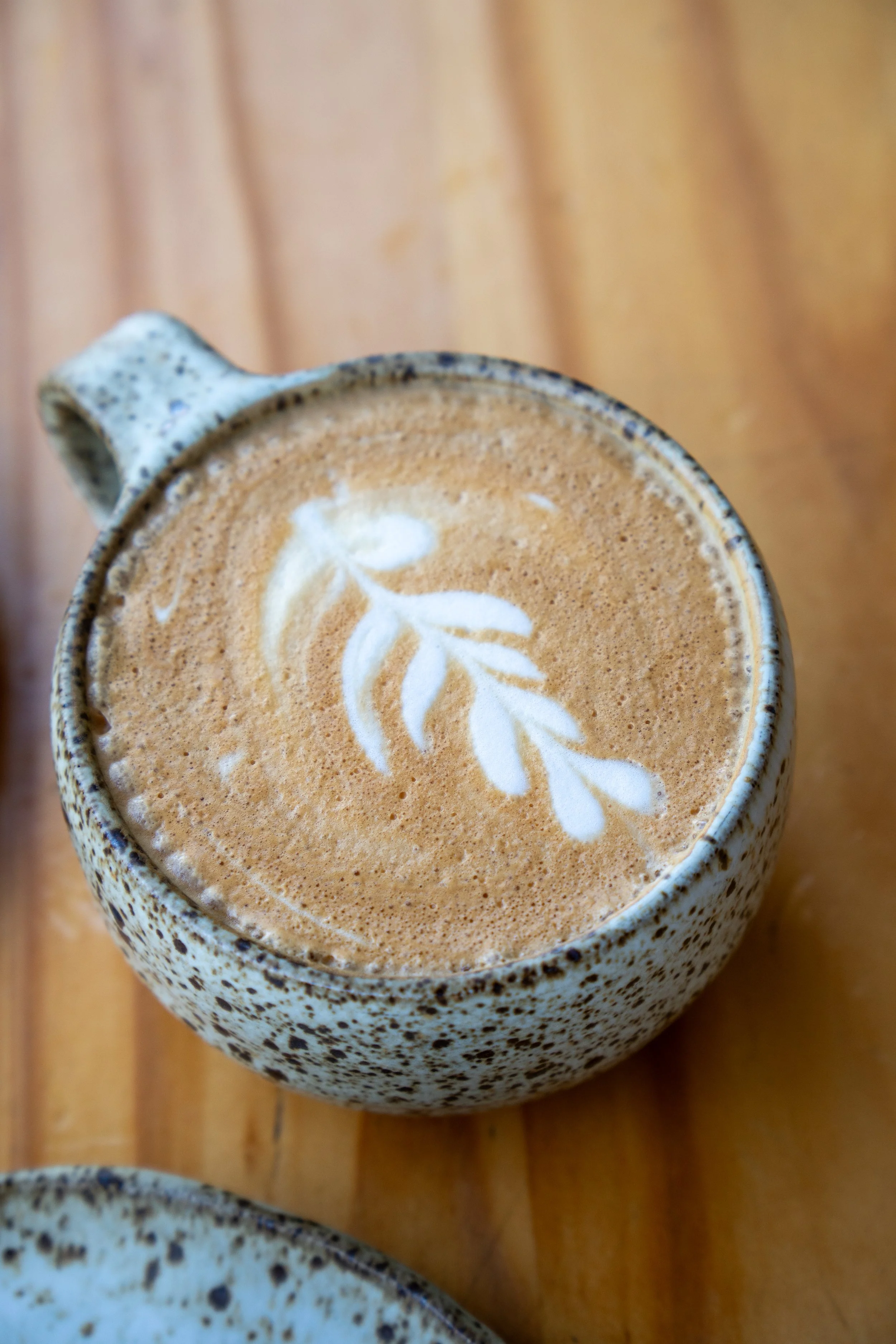 Close-up of a ceramic cup filled with a latte, topped with foam art of a leaf design, placed on a wooden surface.