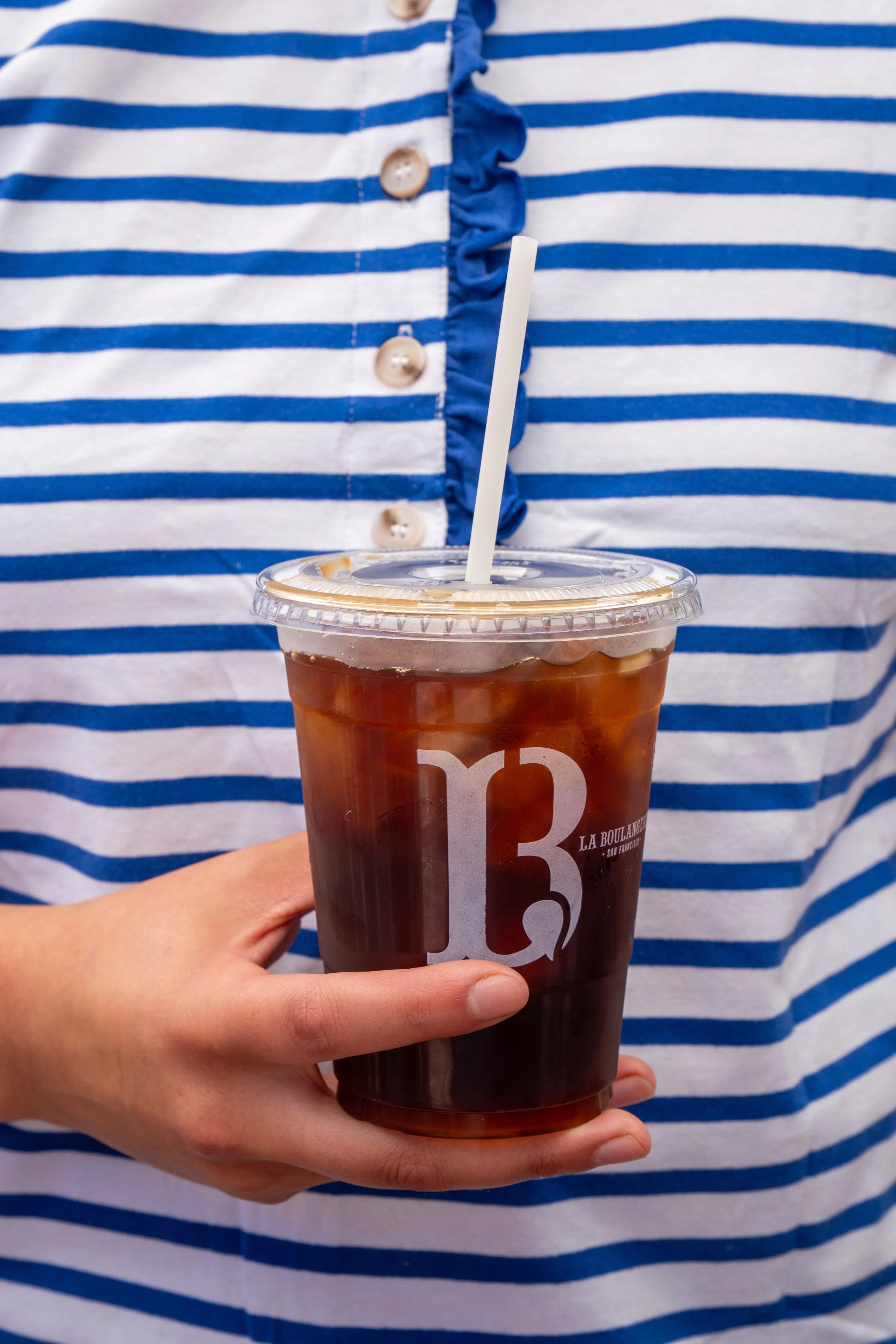 Person wearing a blue and white striped shirt holding a plastic cup filled with dark iced coffee with a straw.