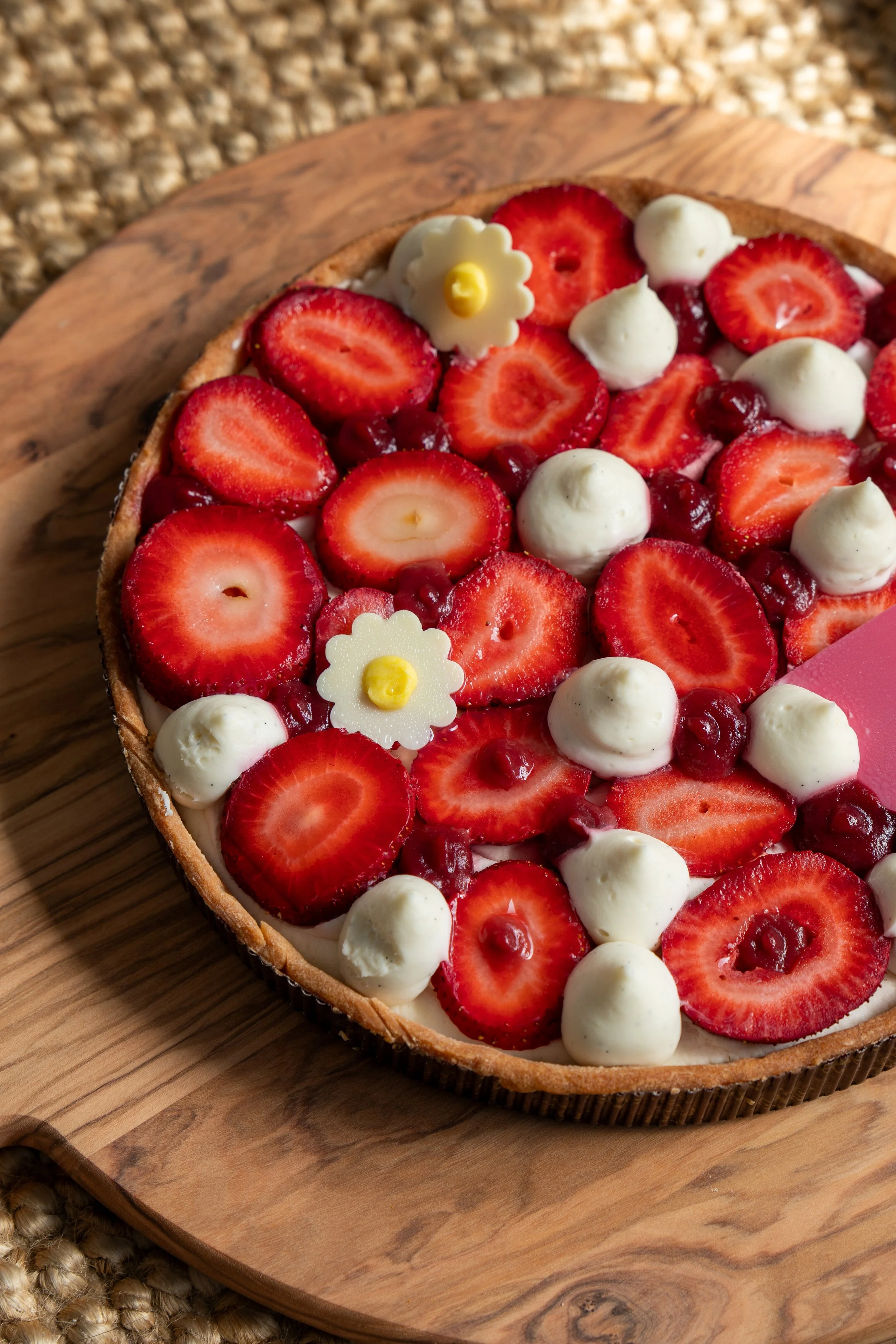 A tart with sliced strawberries, small white dollops of cream, red jelly, and decorative white flowers with yellow centers, sitting on a wooden serving board.