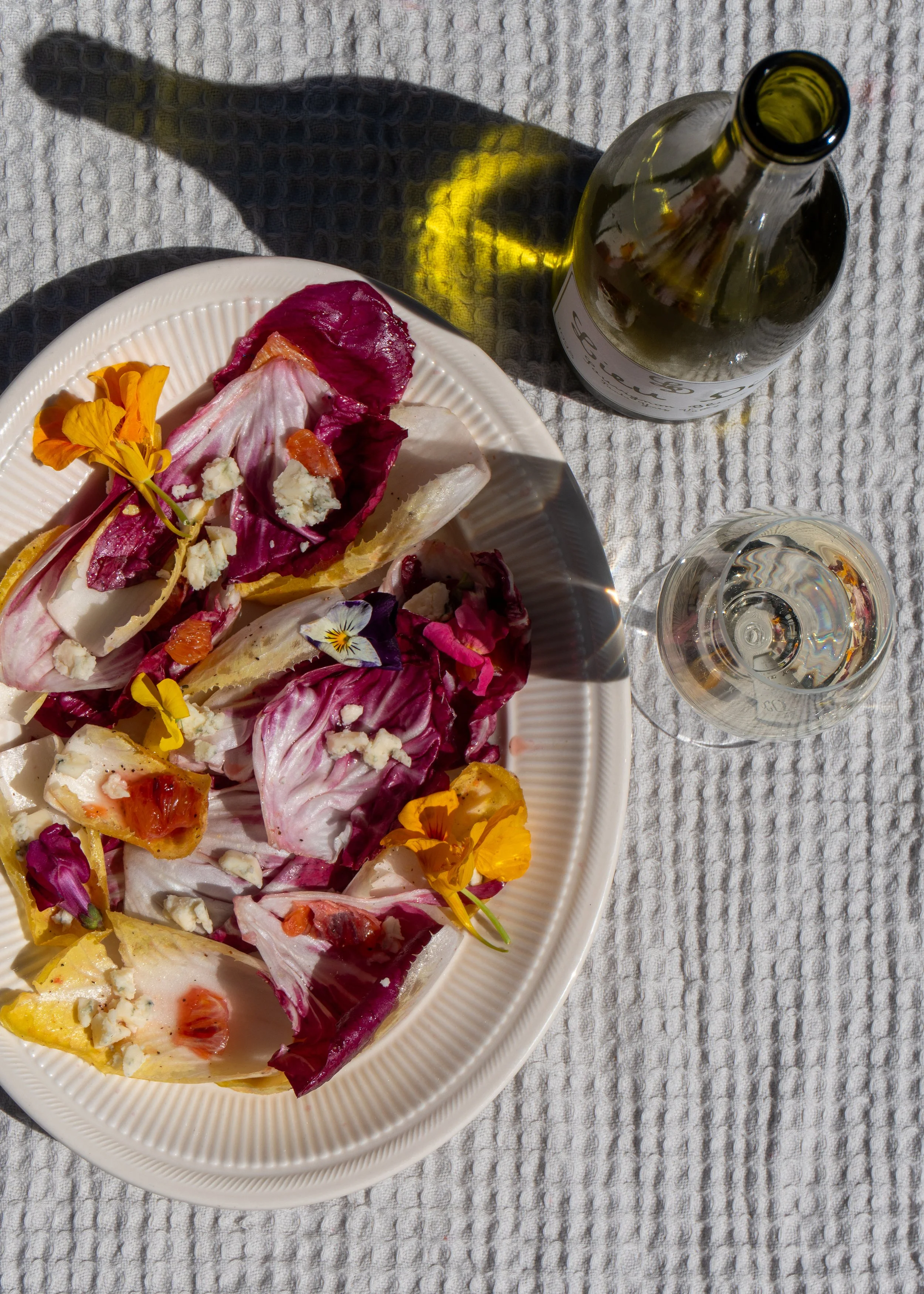 View of a white plate with radicchio and endive salad garnished with edible flowers, cheese, and sliced fruit, on a textured white cloth, with an empty clear wine glass and a glass bottle nearby.