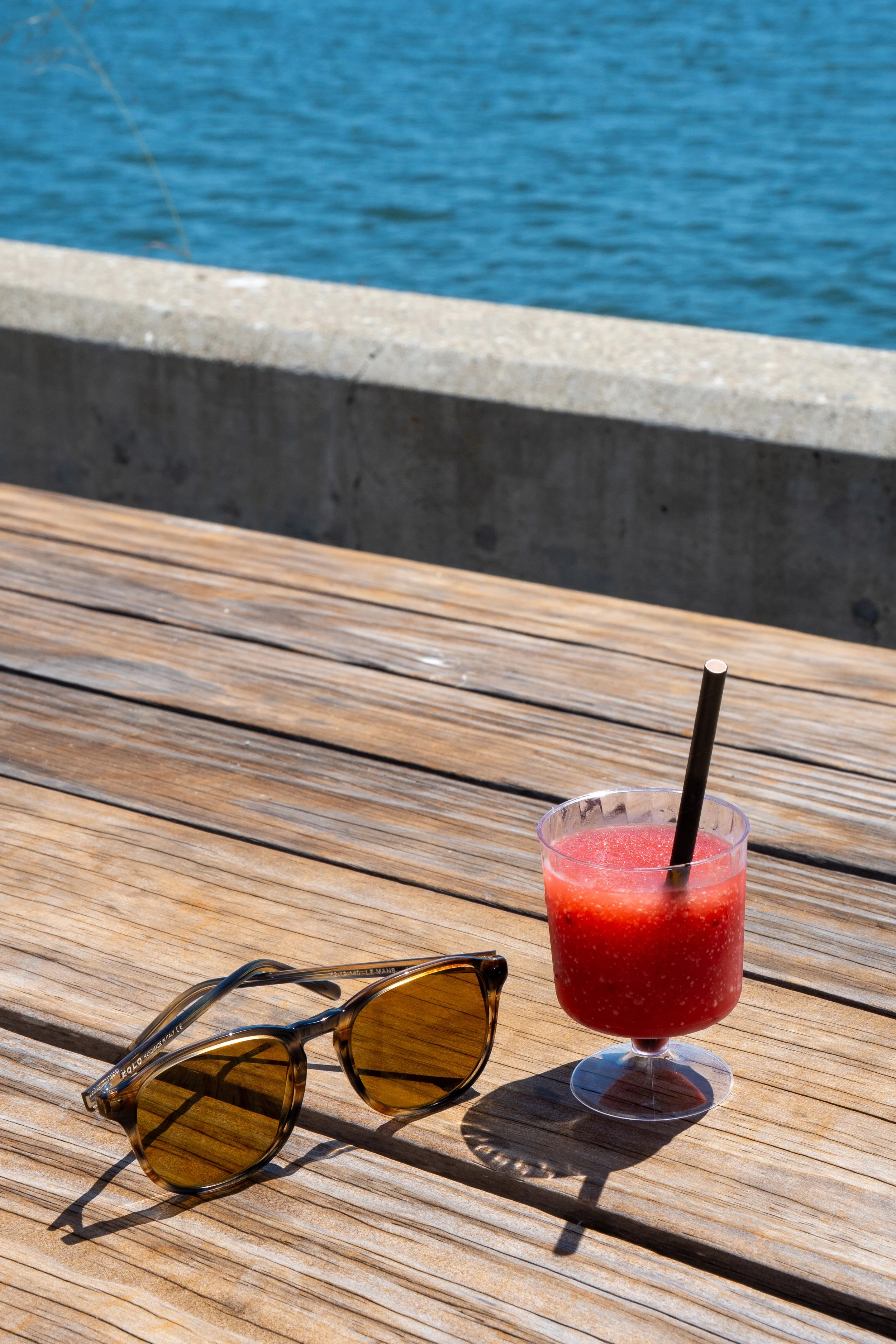 A wooden outdoor table with sunglasses, a pink slushie drink with a black straw, and a waterfront view with blue water and a concrete barrier.