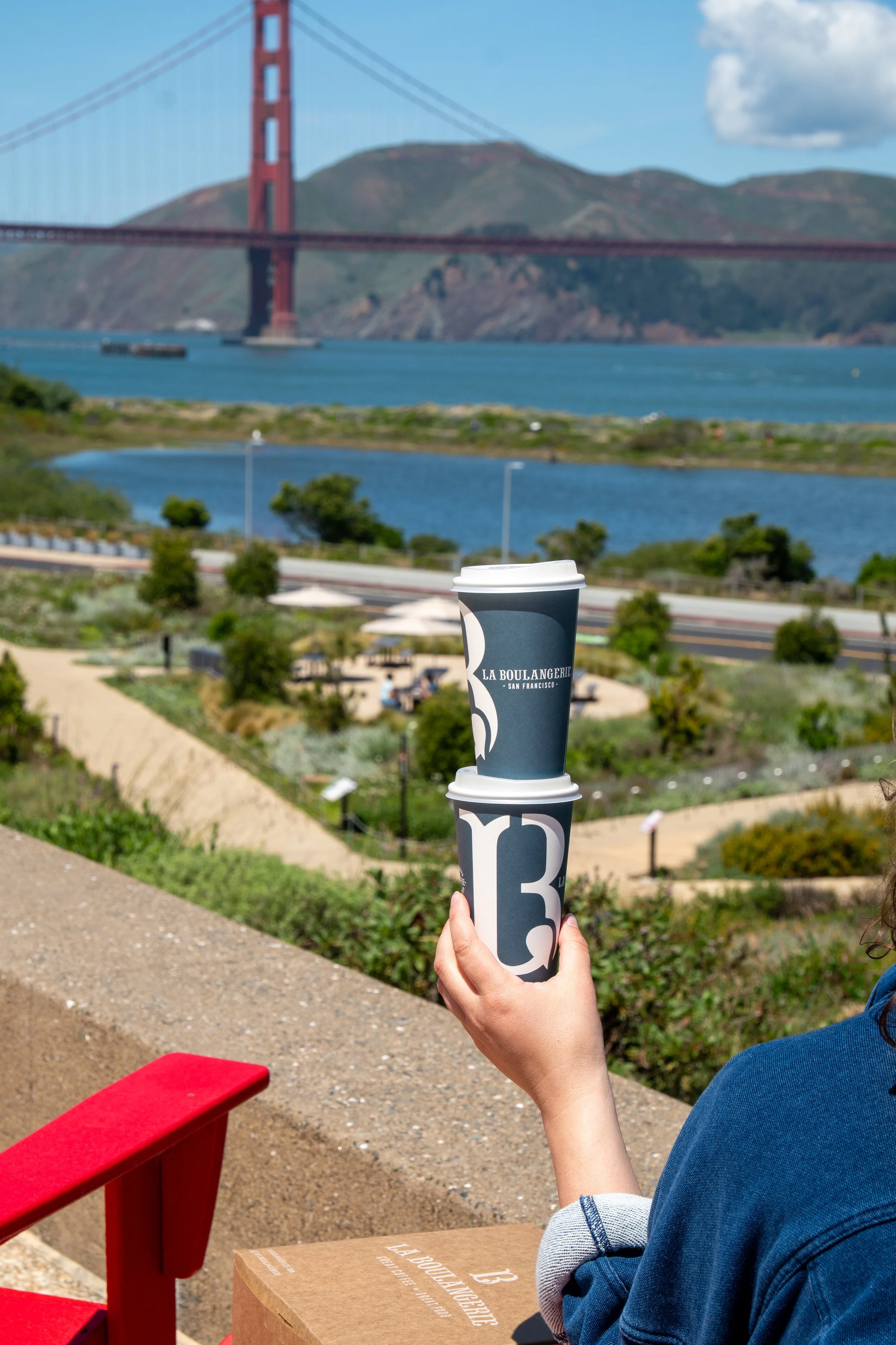 Person holding two stacked coffee cups in front of the Golden Gate Bridge with a view of hills and water in the background.