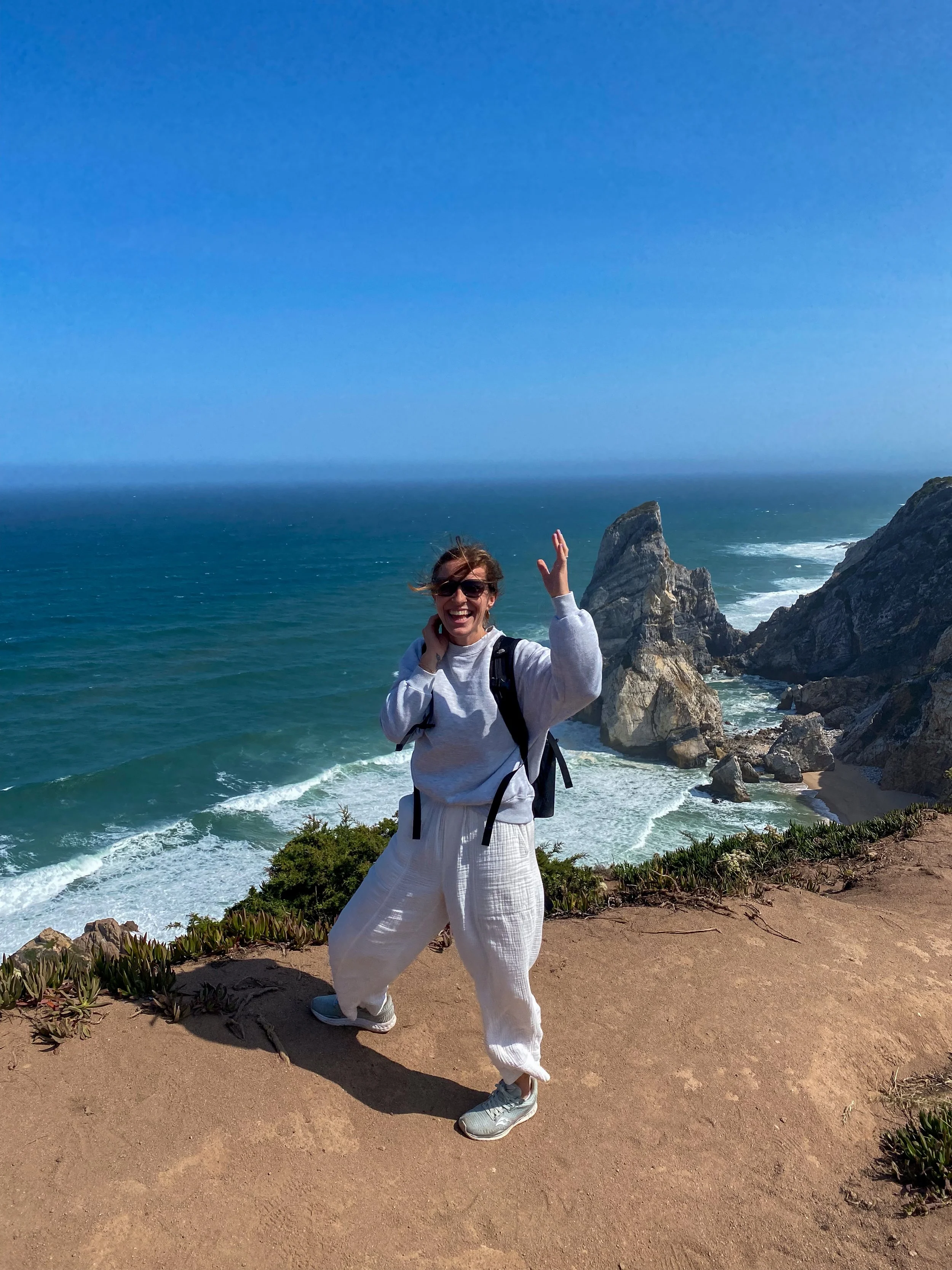A woman smiling and waving at the camera while standing on a dirt path near the ocean, with rocky cliffs and blue sky in the background.