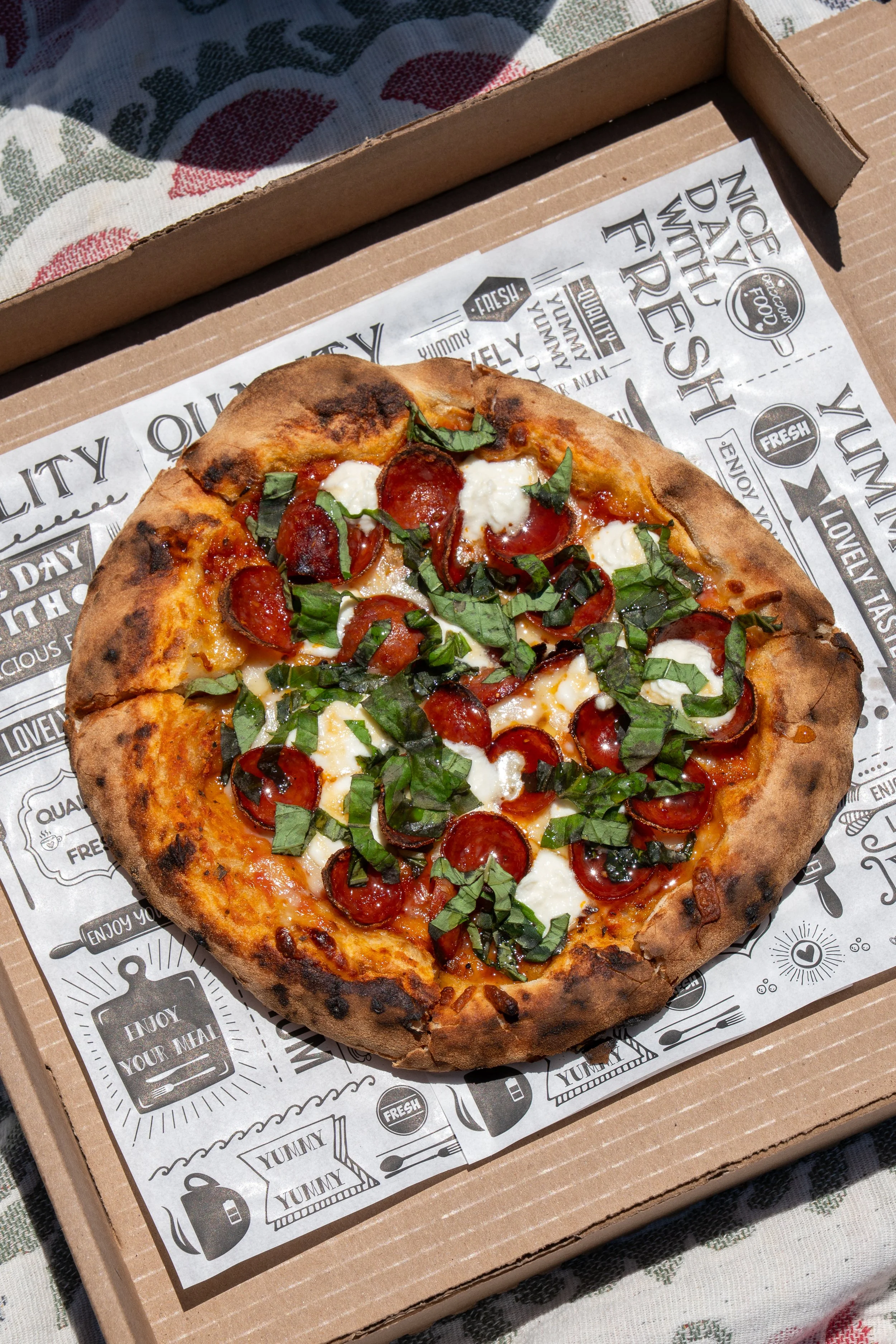A pizza with pepperoni, mozzarella cheese, and chopped basil leaves on a cardboard pizza box lined with printed paper.