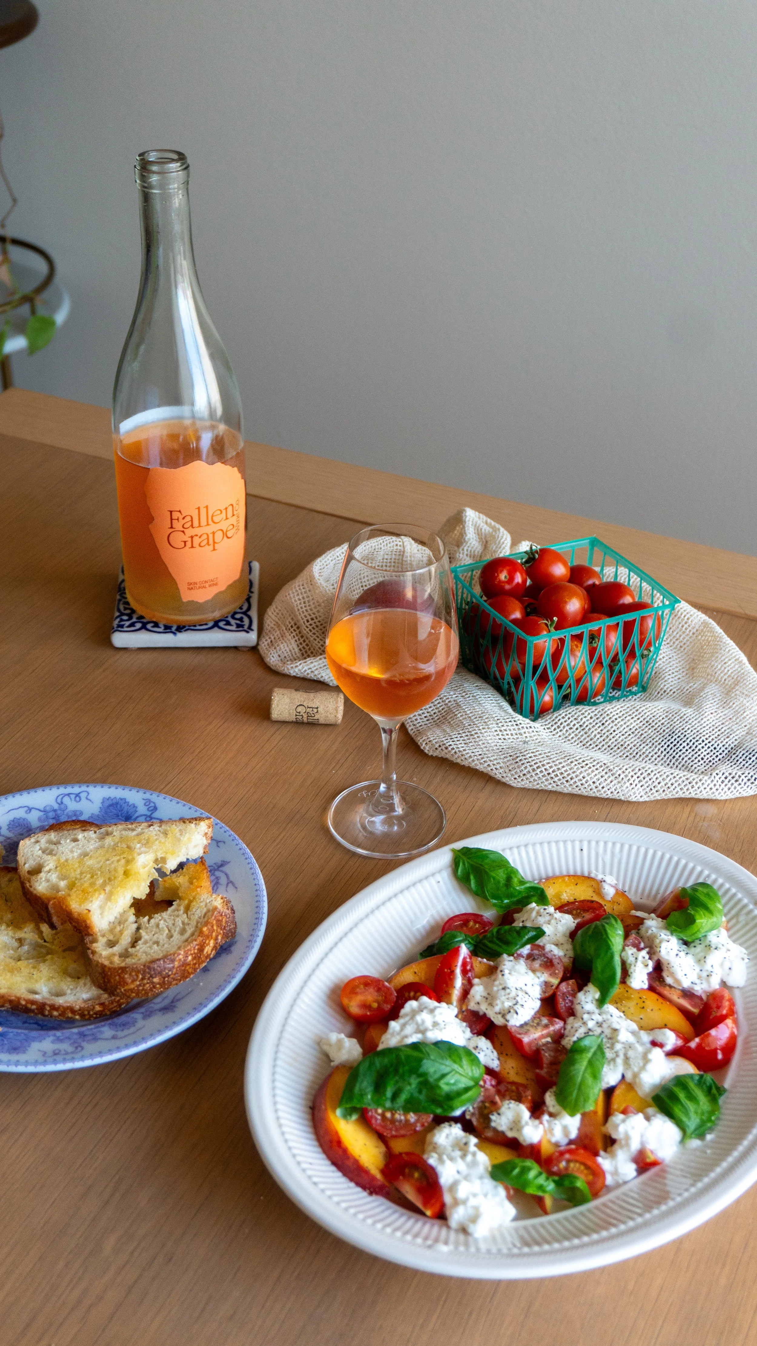 A table with a glass of rosé wine, a bottle of orange wine labeled 'Fallen Grape,' a basket of cherry tomatoes, a plate of pizza with tomatoes and cheese, and slices of bread.