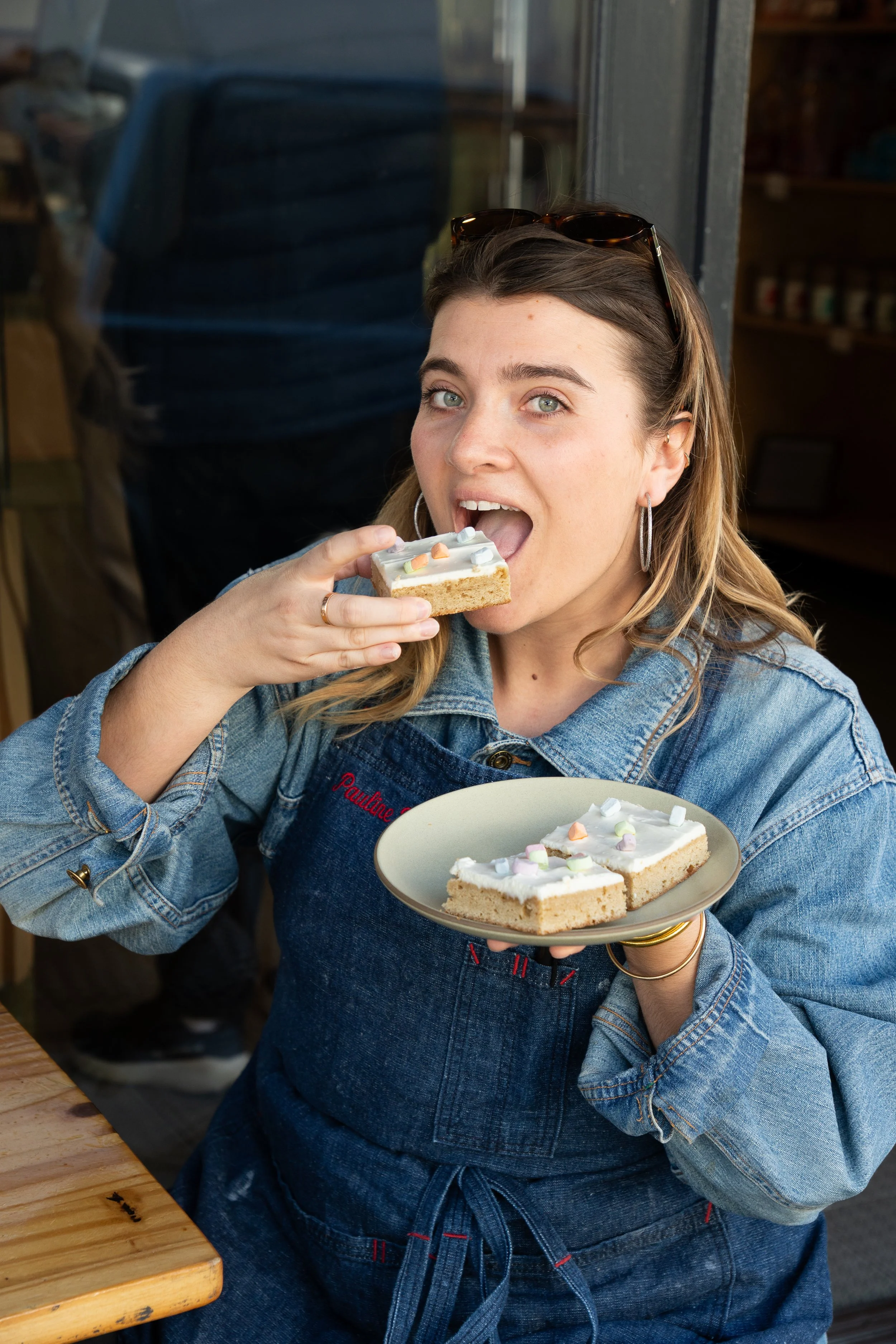 A woman enjoying a slice of cake in a cafe. She is wearing a denim jacket, hoop earrings, and has sunglasses on her head. She holds a plate with more cake slices.