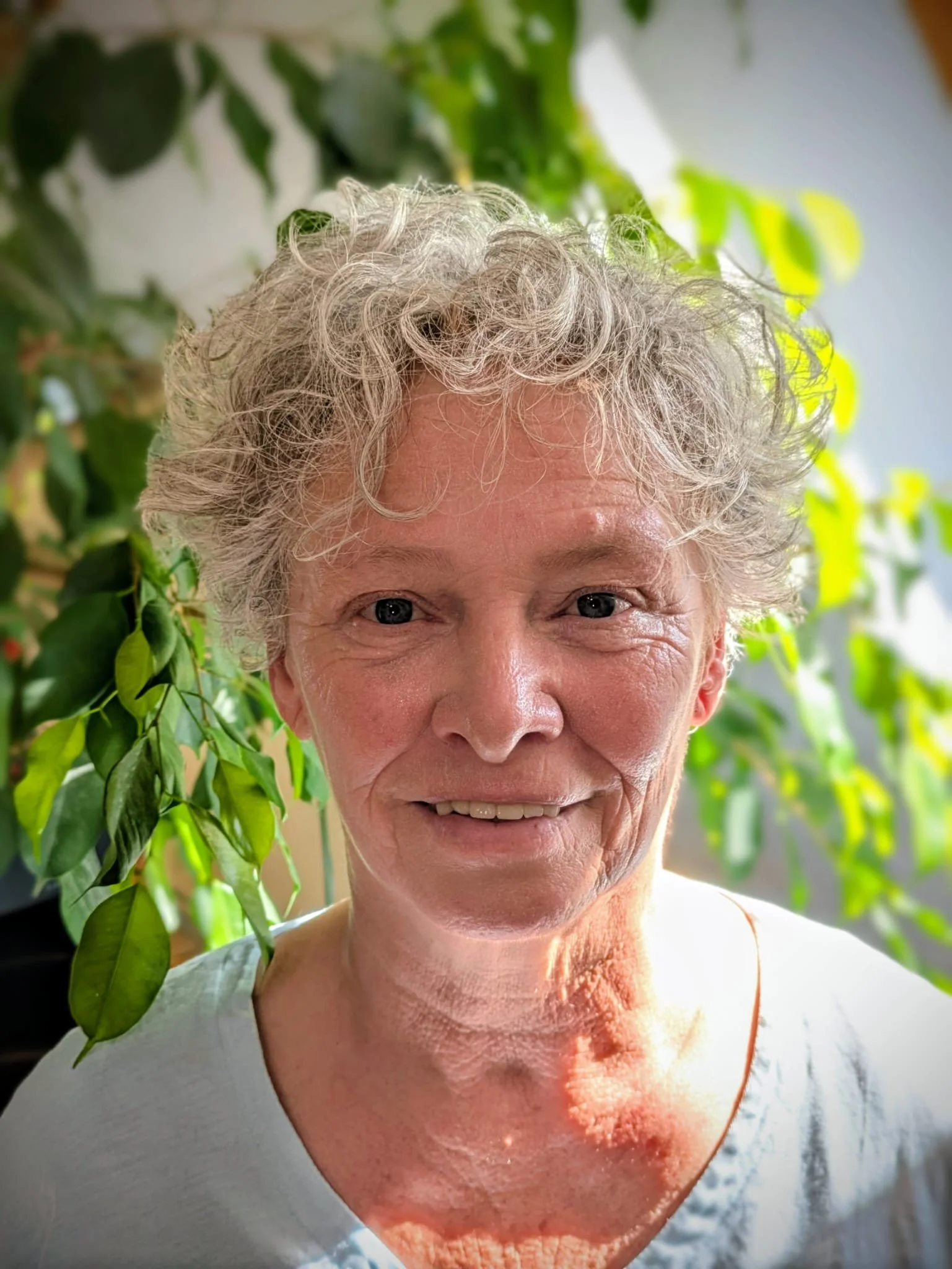 Nahaufnahme einer älteren Frau mit lockigem, grauem Haar, vor einem Hintergrund aus grünen Pflanzen, lächelnd.