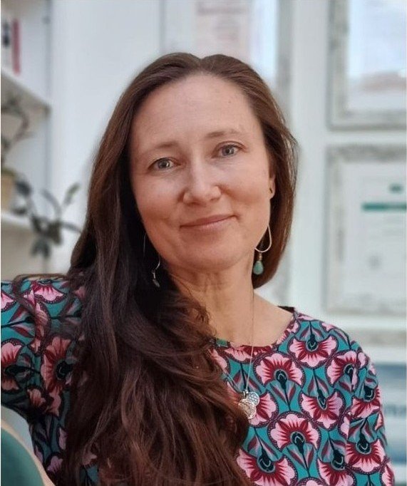 Eine Frau mit langen braunen Haaren, die ein farbenfrohes, blumenmusterndes Kleid trägt, sitzt vor einer neutralen, hellen Hintergrund mit einigen Bildern oder Dokumenten an der Wand.