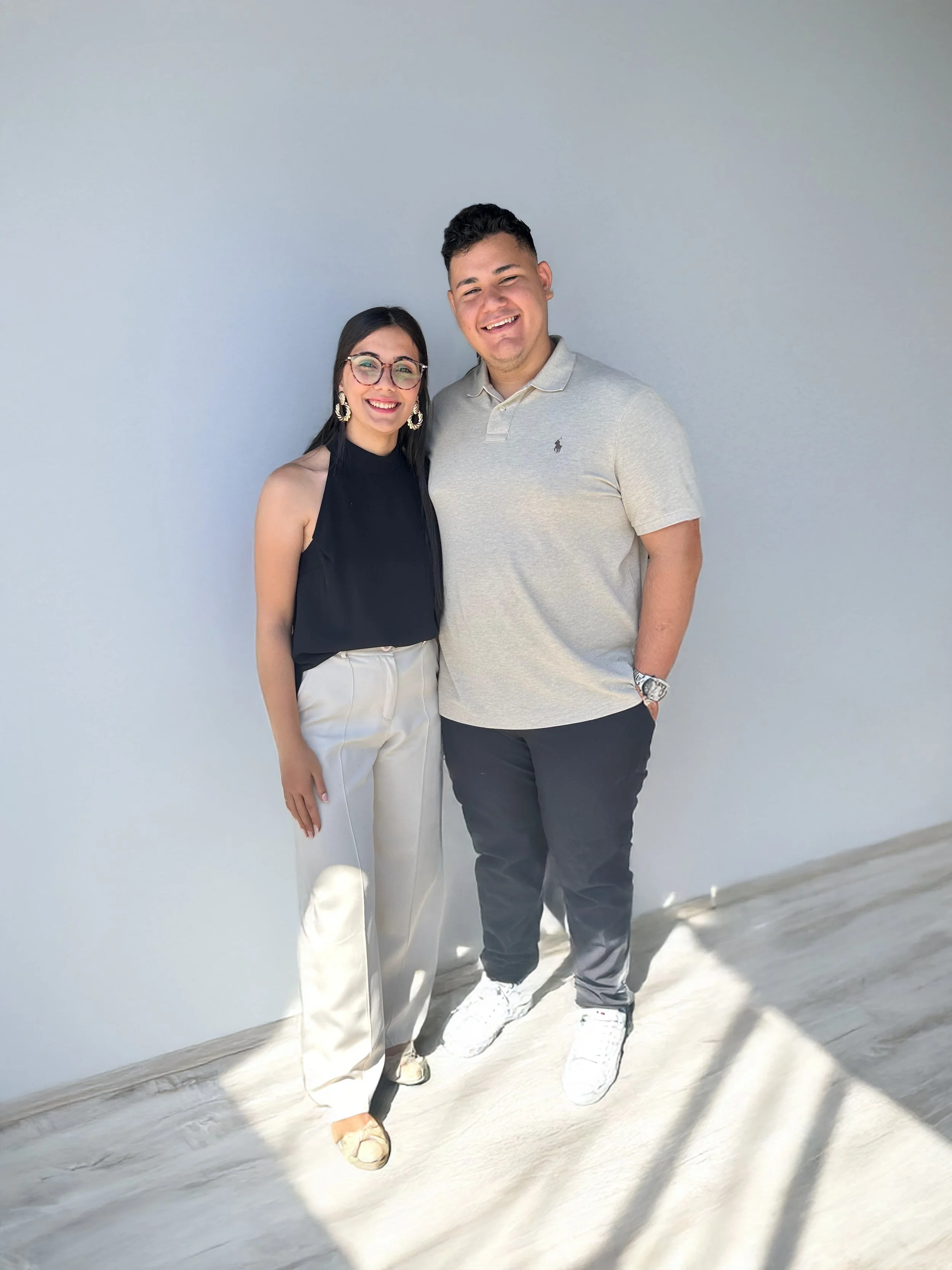 Dos personas sonrientes posando frente a una pared blanca. Una mujer con gafas y blusa negra y un hombre con polo gris claro.