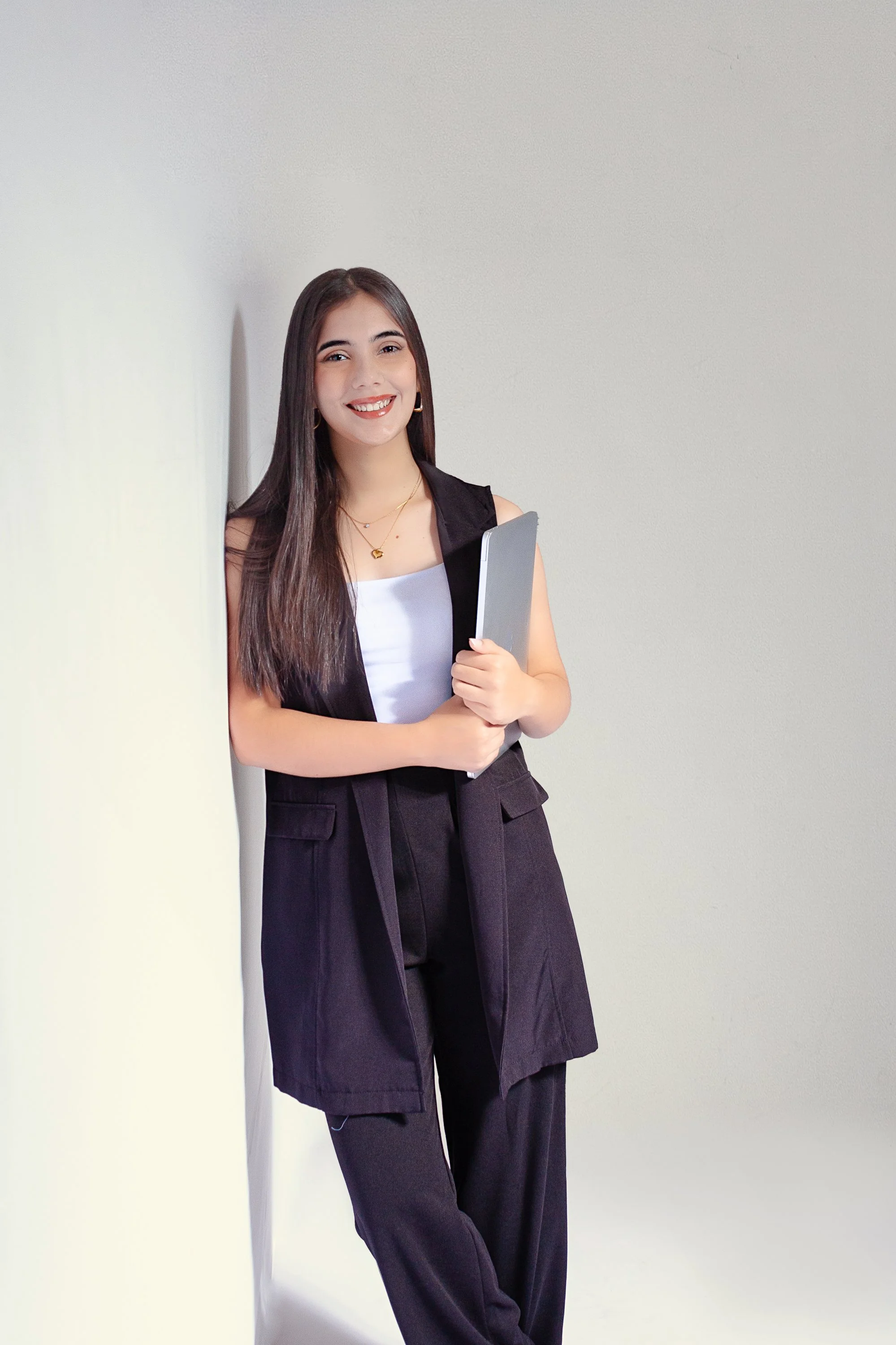Mujer joven sonriendo, vestida de negro, sostiene una laptop y está apoyada contra una pared.