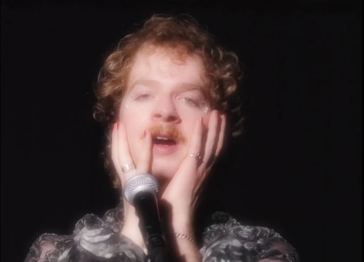 A woman with curly hair and a floral dress appears to be in the middle of an emotional performance, with her hands on her face, near a microphone.