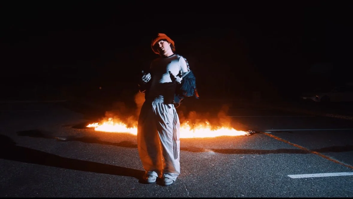 Person standing in front of a fire on a dark street at night, wearing casual clothing and a beanie.