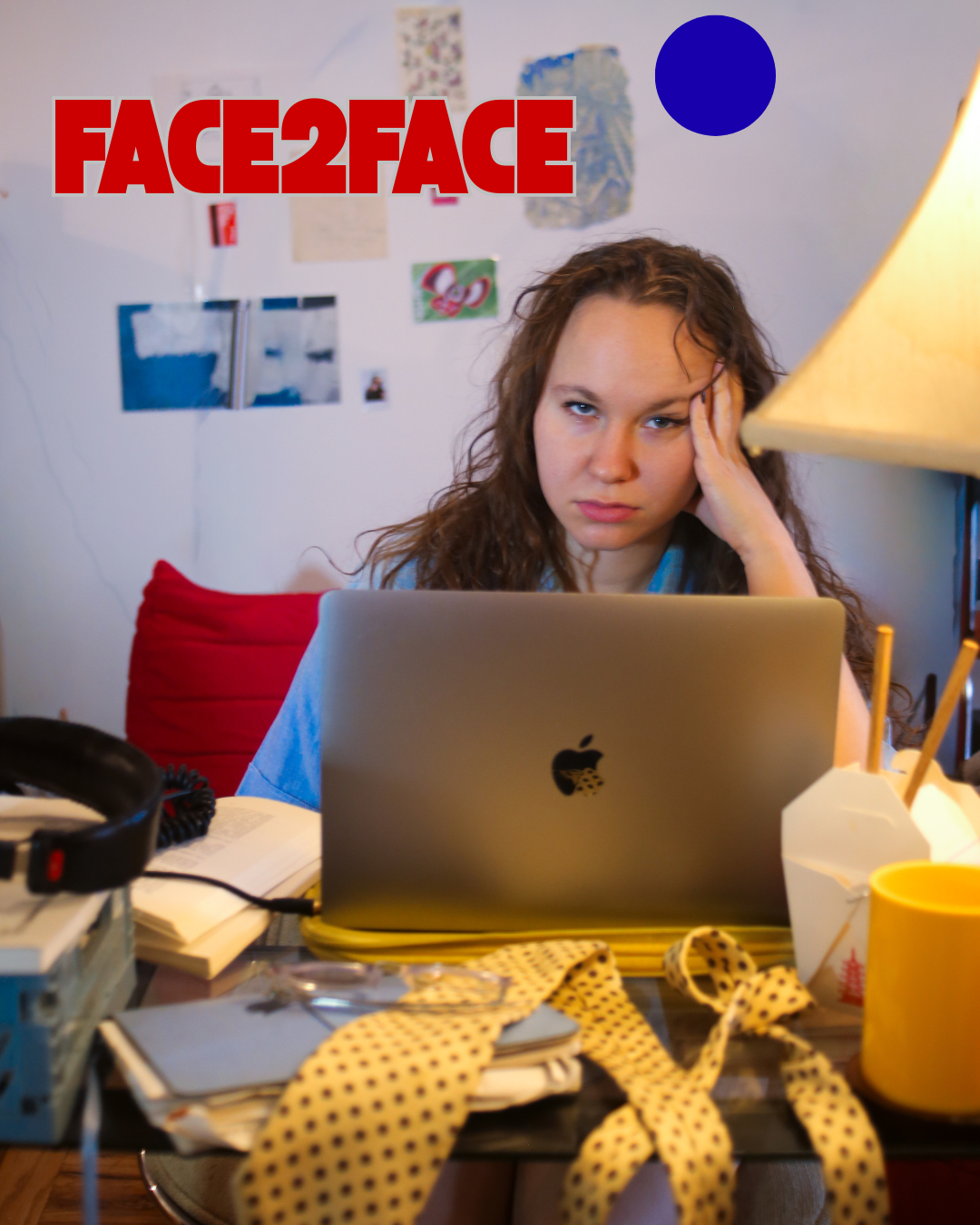 A young woman with curly brown hair working on a laptop at a cluttered desk, with a stressed expression, resting her forehead on her hand, in a room with a white wall decorated with pictures and notes. The desk has various items including papers, a yellow polka dot ribbon, a yellow cup, and a lamp.